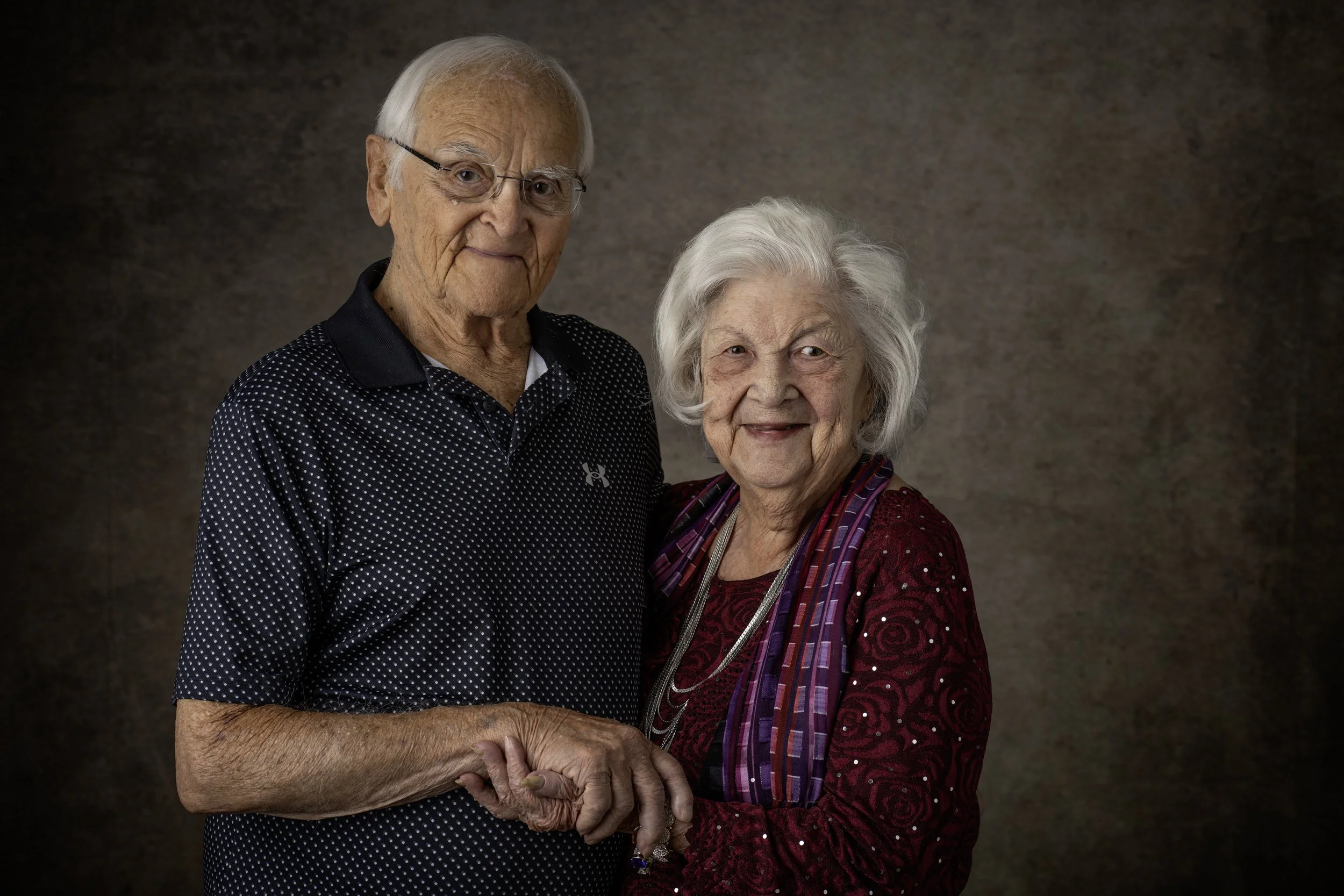 An elderly man and woman holding hands and smiling at the camera against a brown background.