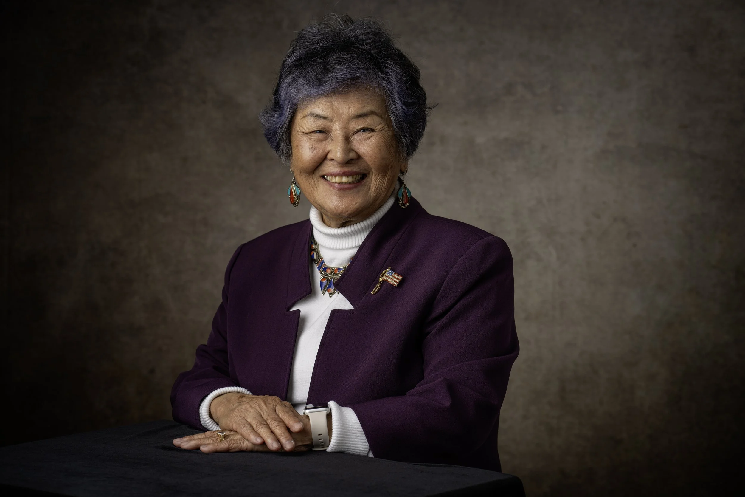 A smiling elderly Asian woman with short gray hair, wearing a maroon blazer, a white turtleneck, colorful jewelry, and an American flag pin, sitting at a dark table against a textured neutral background.