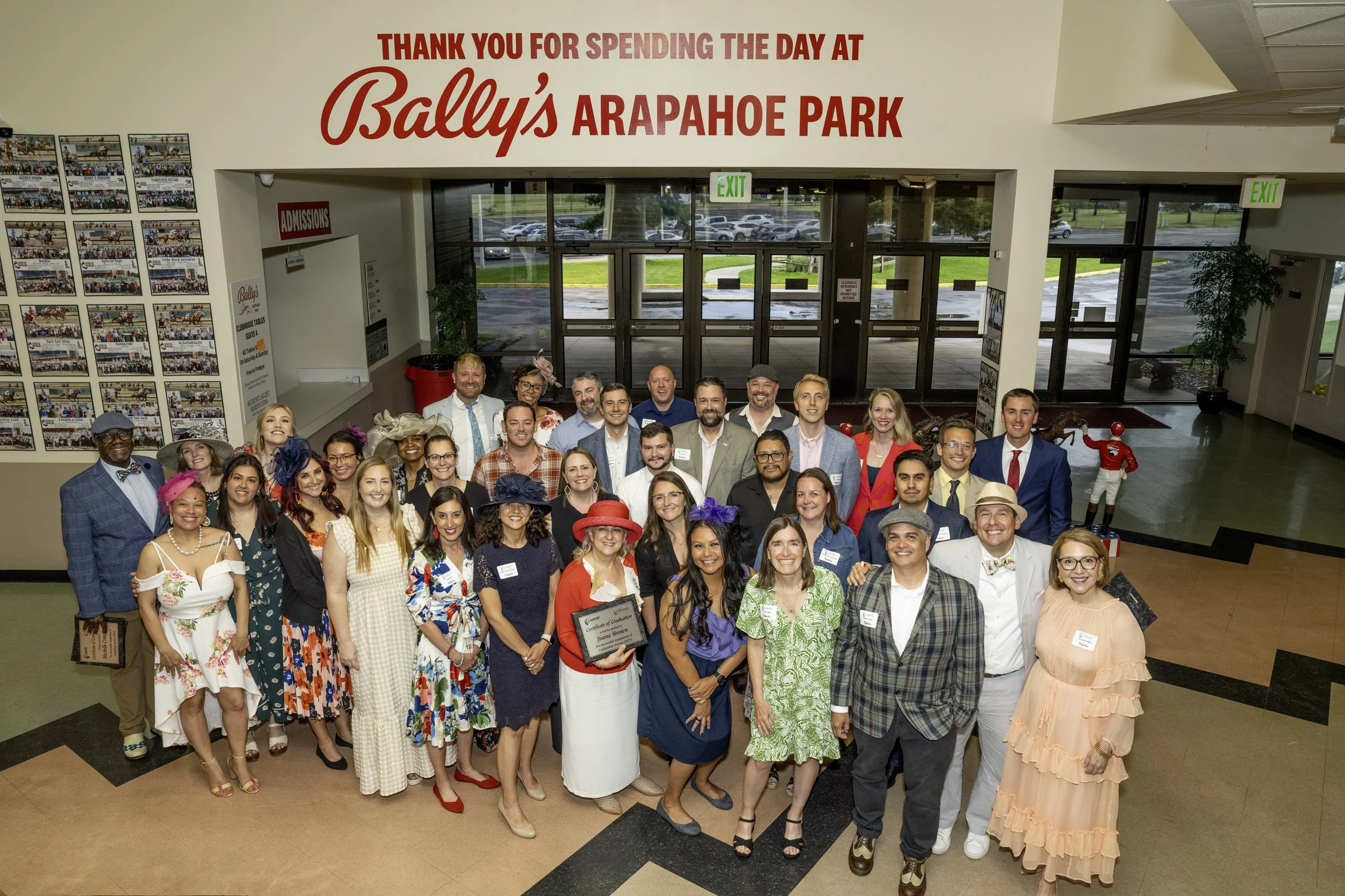 Group photo of people at Bally's Arapahoe Park, with a banner thanking guests for their visit and a display of photos on the wall. The group includes men and women dressed in colorful and formal attire, some wearing hats, standing in the lobby area w