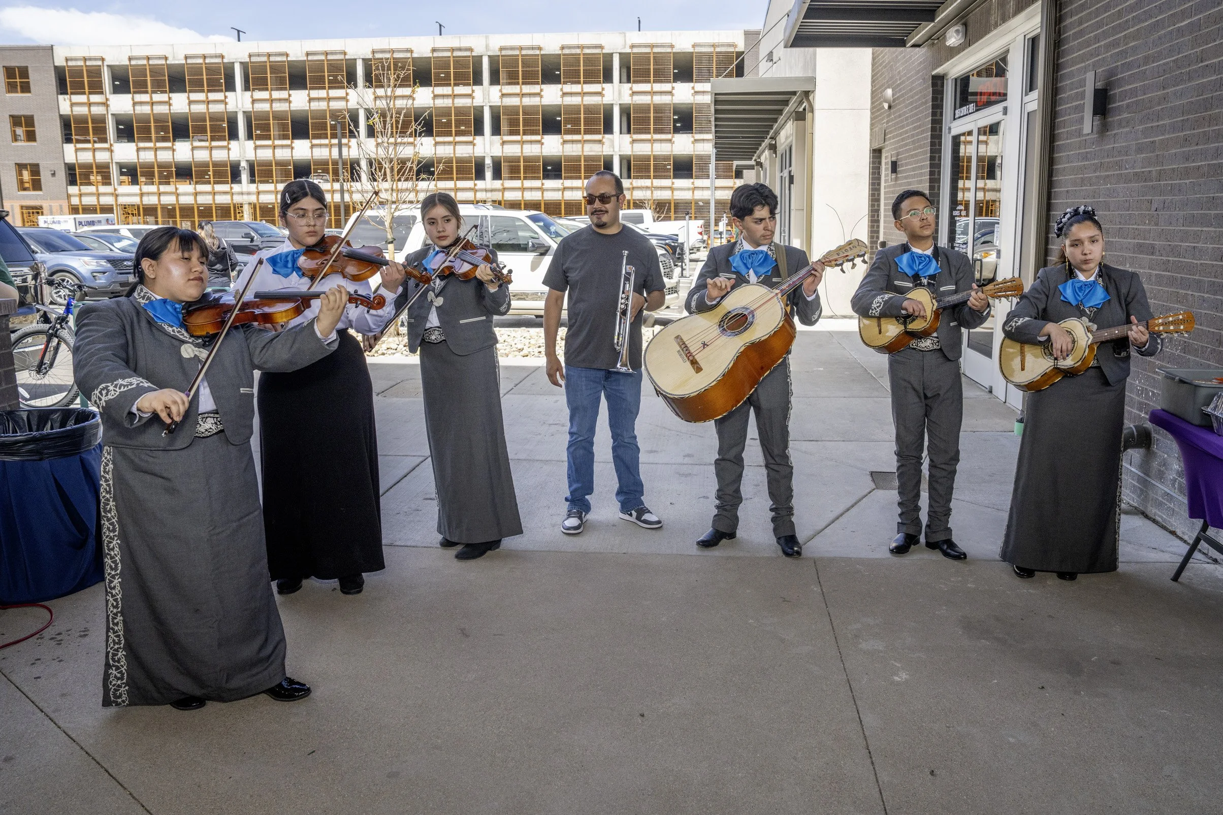 A group of musicians performing outdoors, including young girls in traditional Mexican attire playing violins, a man holding a trumpet, and others playing guitars, standing on a sidewalk in front of a building, with cars parked behind them.