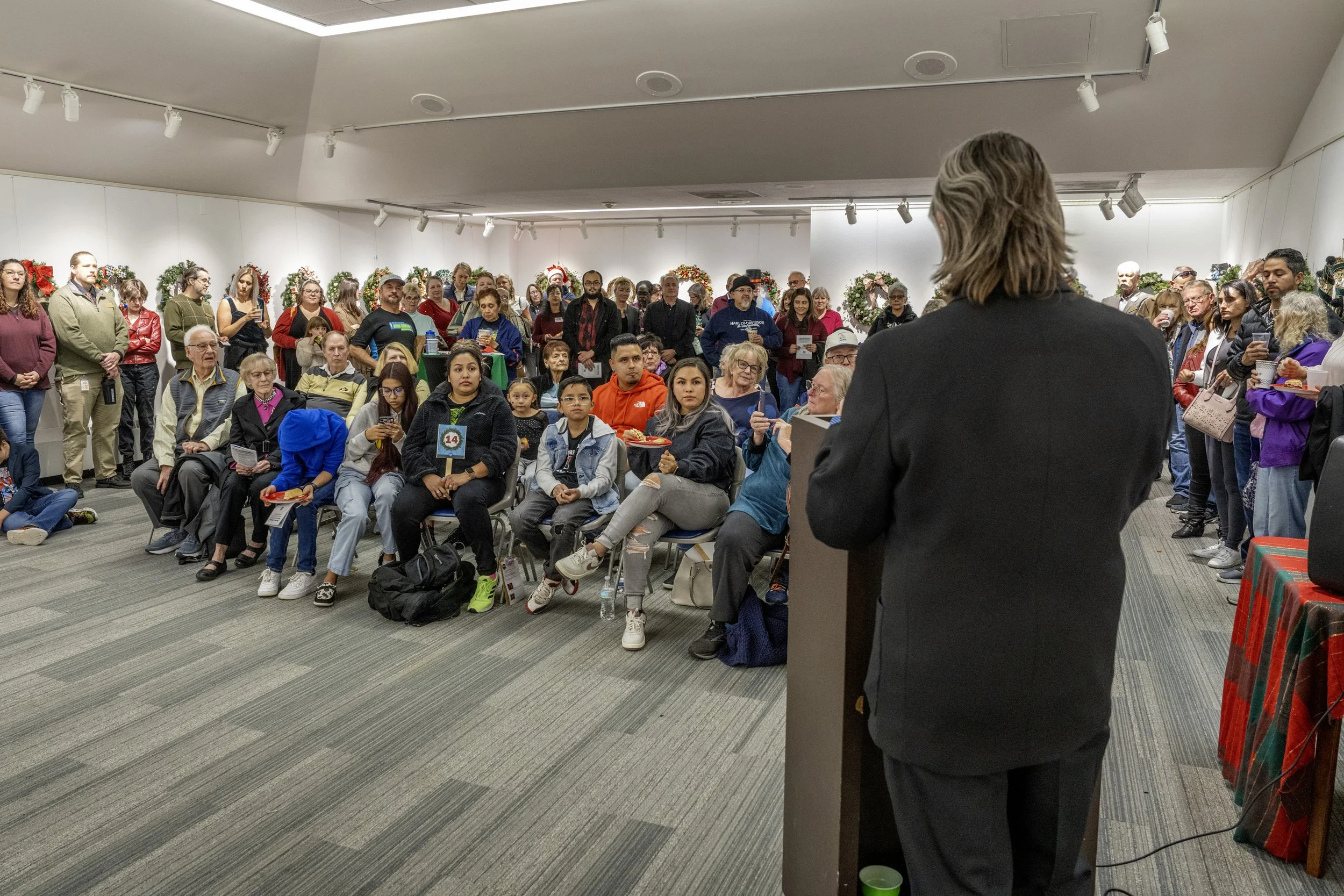 A speaker in a black suit addressing a diverse group of people at an indoor event decorated with holiday wreaths and garlands, some audience members seated, others standing.