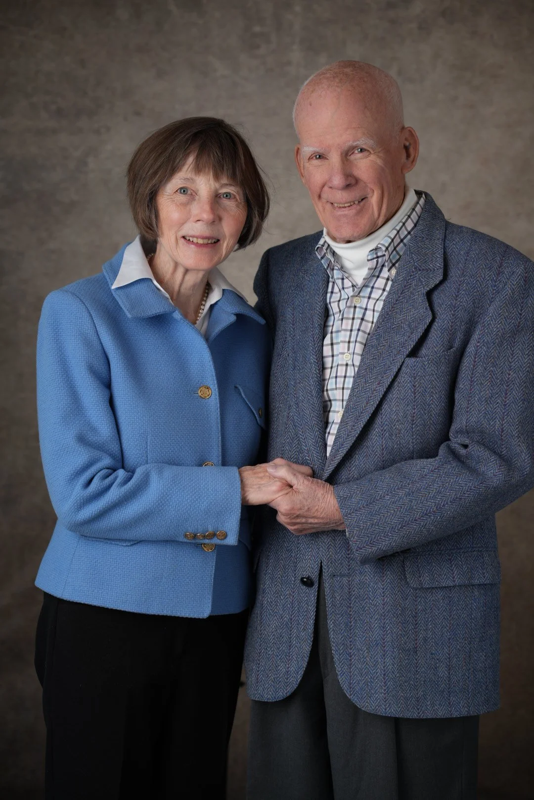 An elderly couple holding hands and smiling at the camera, standing against a neutral background.