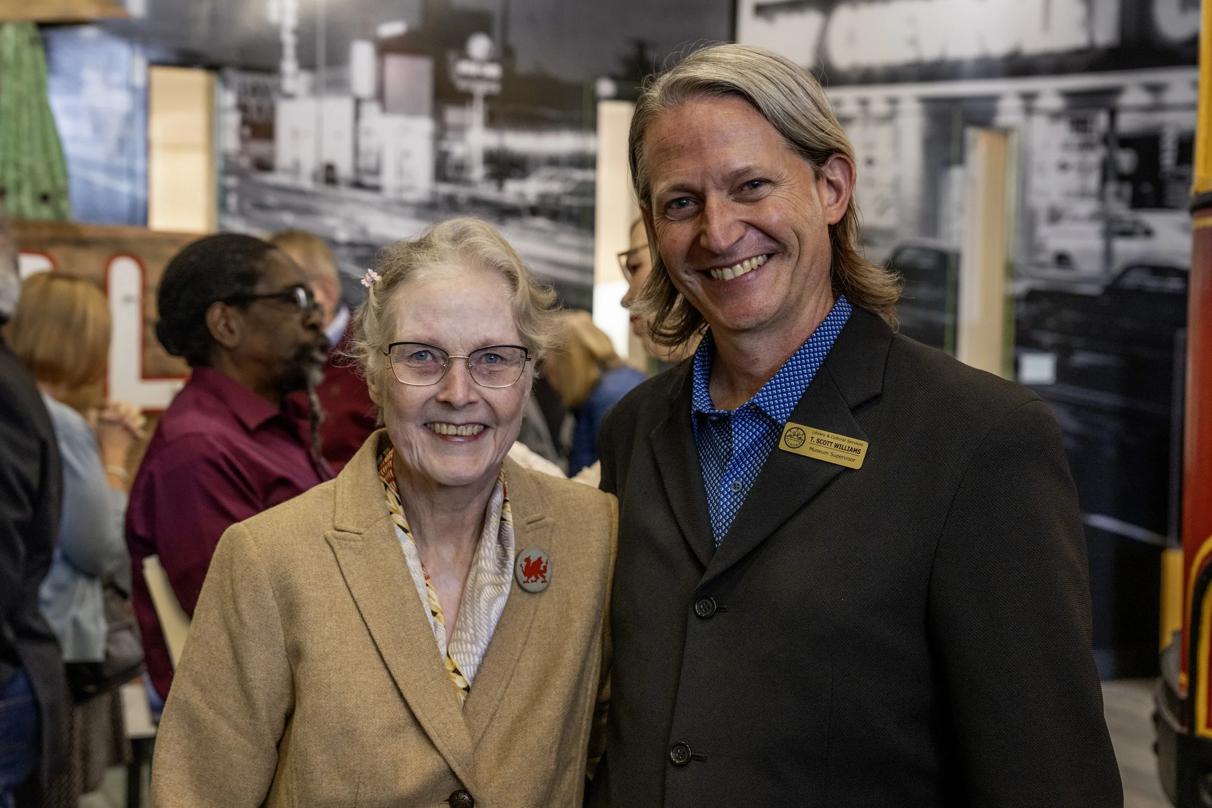 An older woman with gray hair, glasses, and a tan blazer standing next to a smiling middle-aged man with shoulder-length blond hair, wearing a dark blazer and a name tag, at an event with a crowd and artwork displayed in the background.