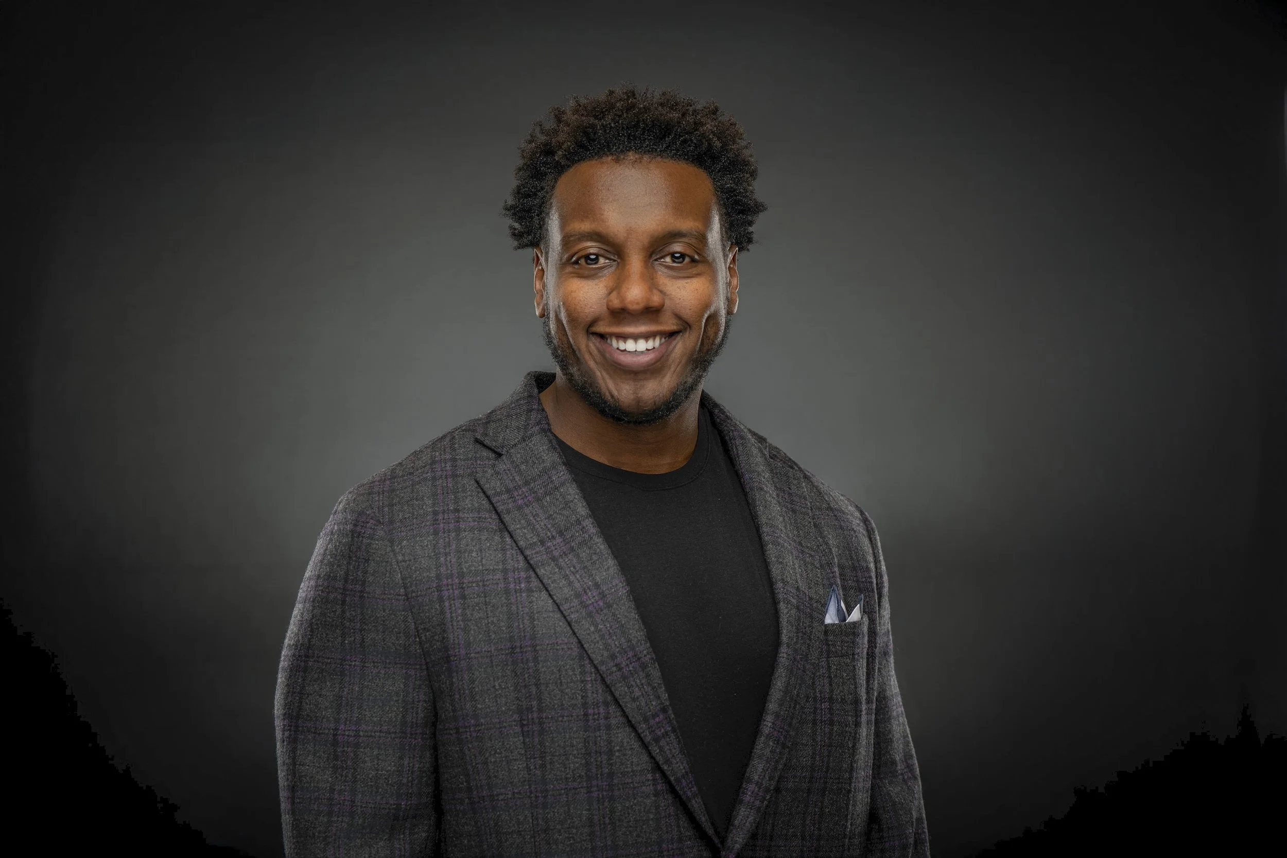 A young man with dark skin, curly hair, and a beard smiling, dressed in a black shirt and a gray plaid blazer, against a dark gradient background.