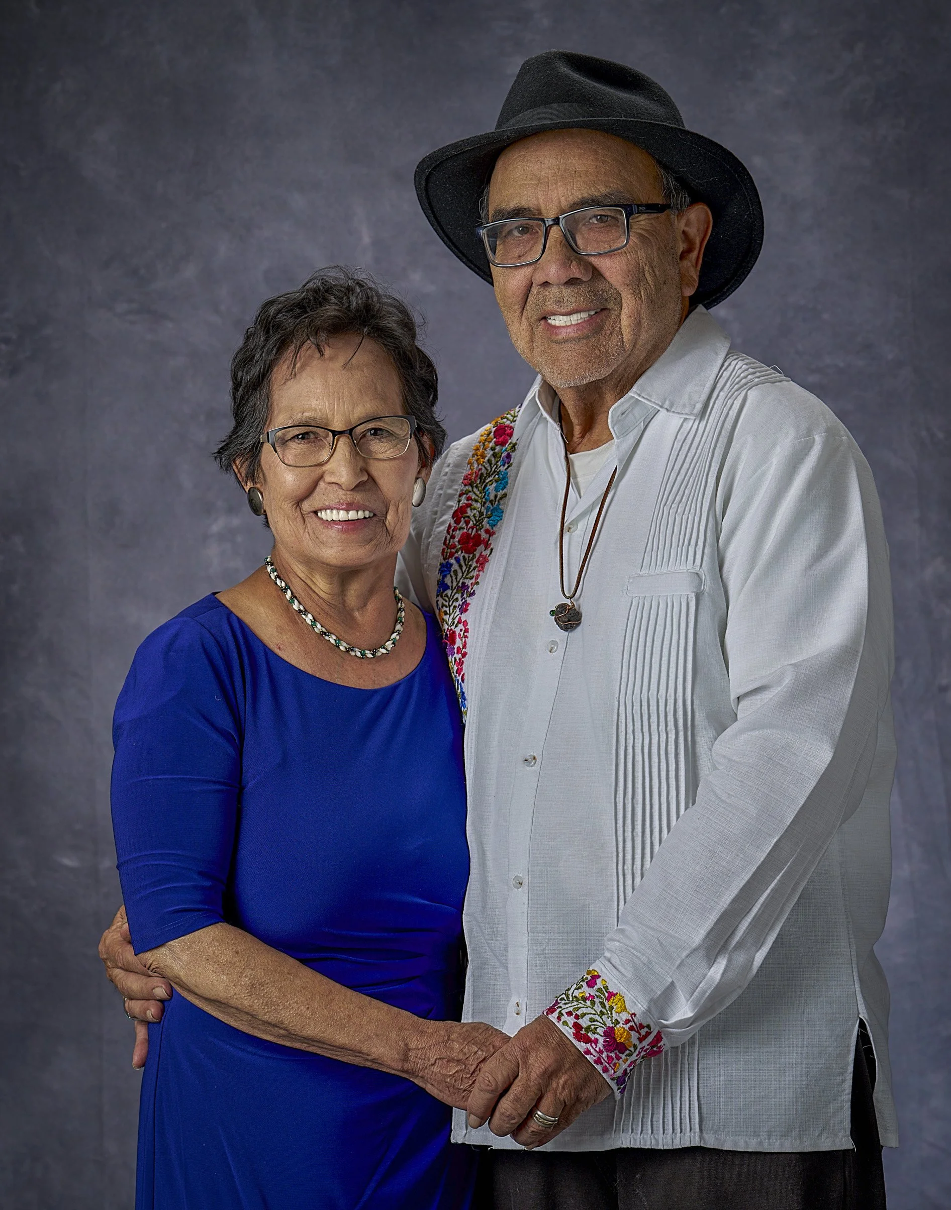 A senior couple posing together, the woman in a blue dress and the man in a white shirt with embroidered cuffs, both wearing glasses and holding hands against a gray textured background.