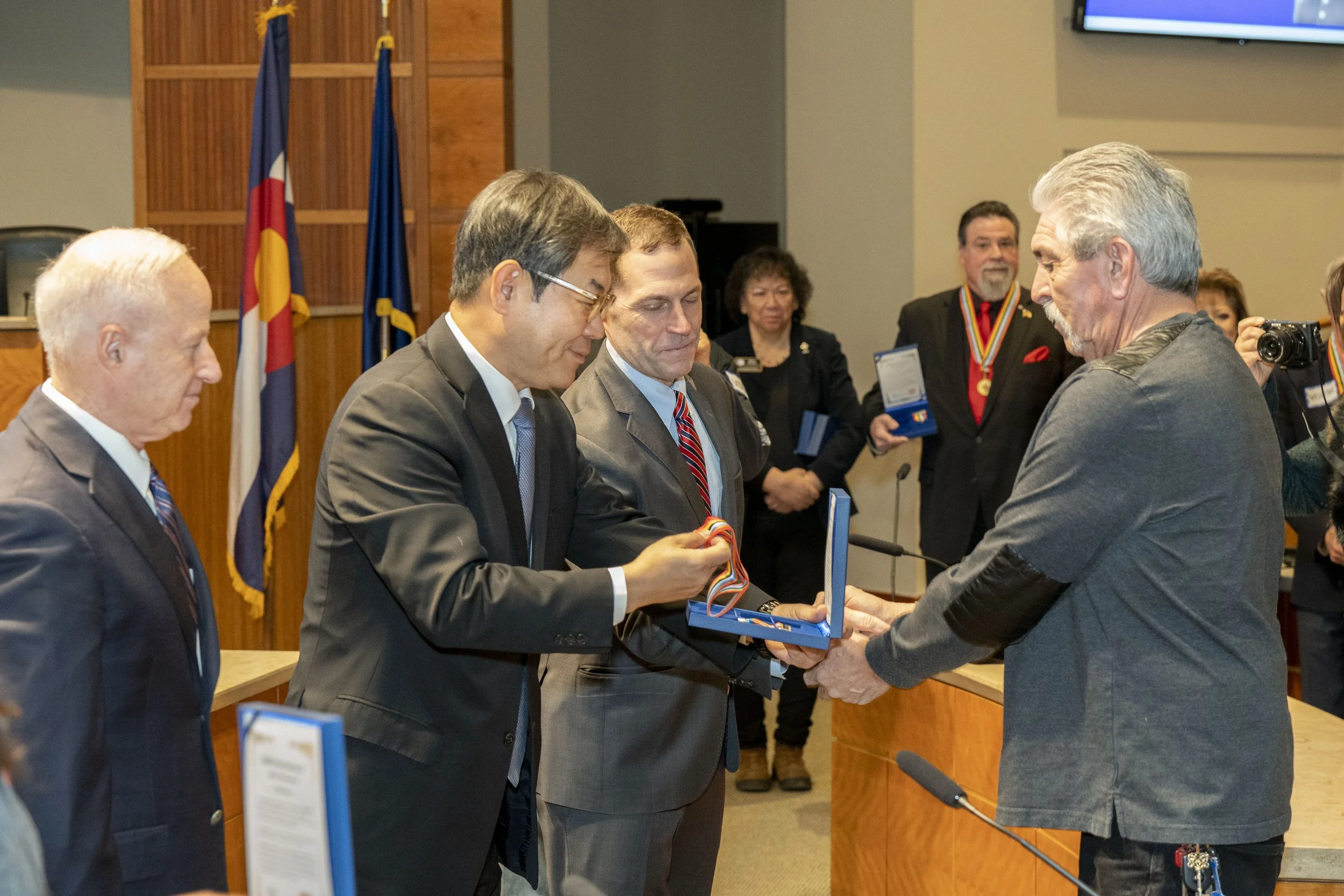A man with gray hair and a beard receives a medal from another man during an award ceremony in a conference room. Several people, some holding medals and plaques, watch the event. Flags in the background include the Colorado state flag and the U.S. f