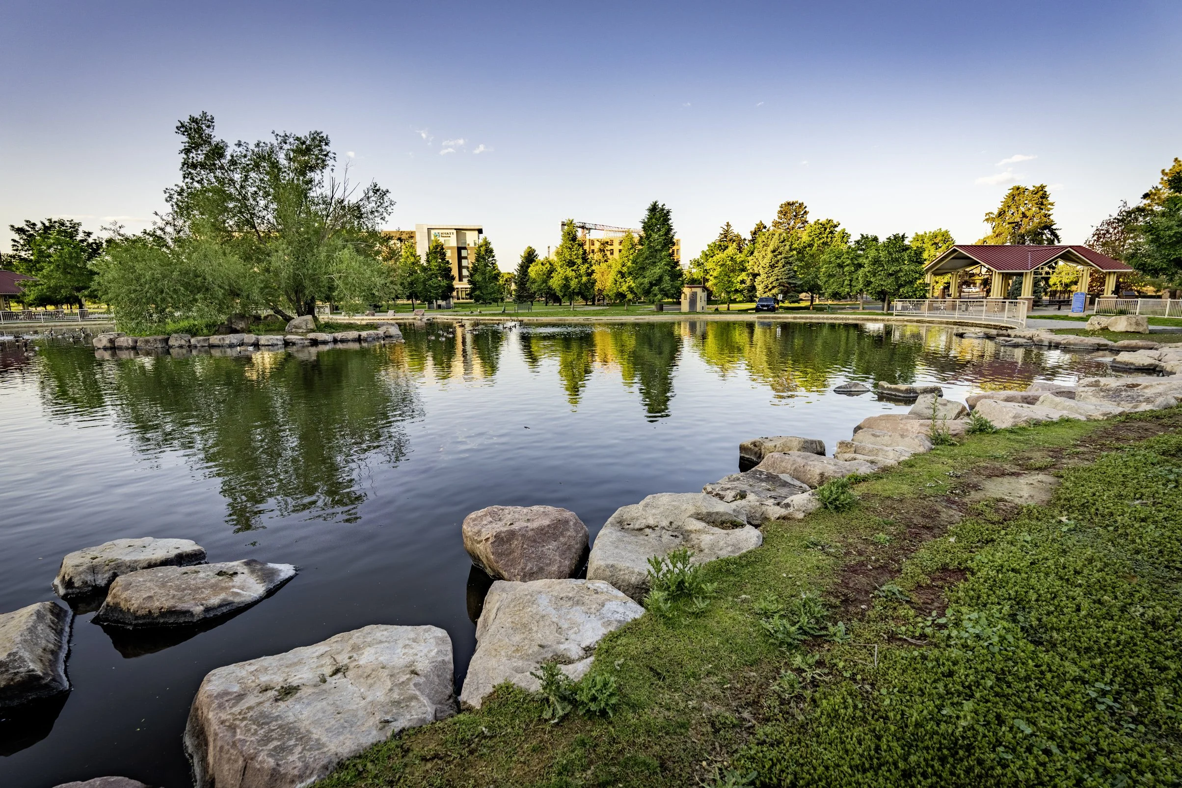 A scenic park with a calm pond reflecting the trees and structures around it, a rocky shoreline, green grass, and a clear sky.