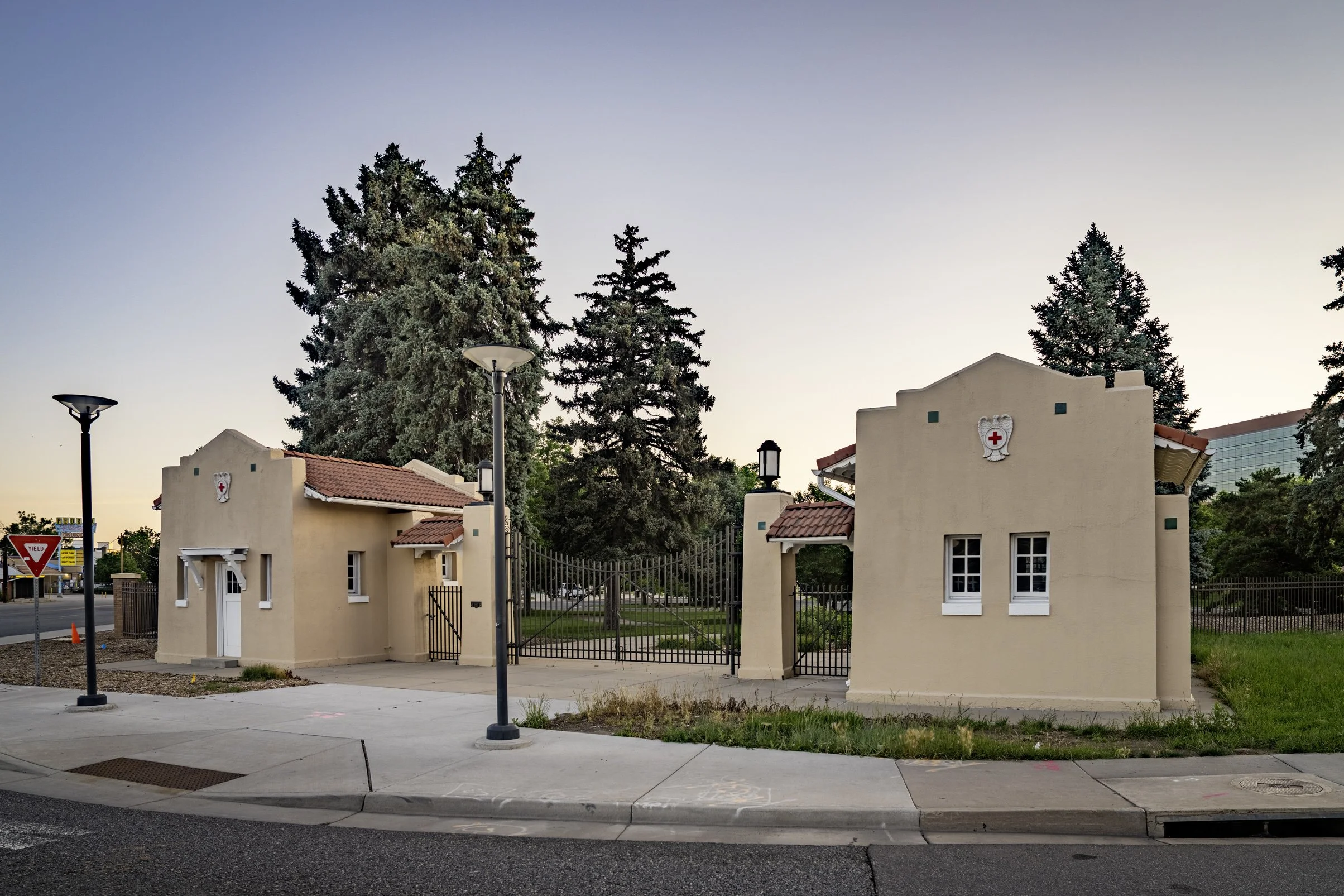Two beige stucco buildings with red tile roofs and a mural of a red cross emblem, surrounded by trees, with a sidewalk and street in the foreground at dusk.