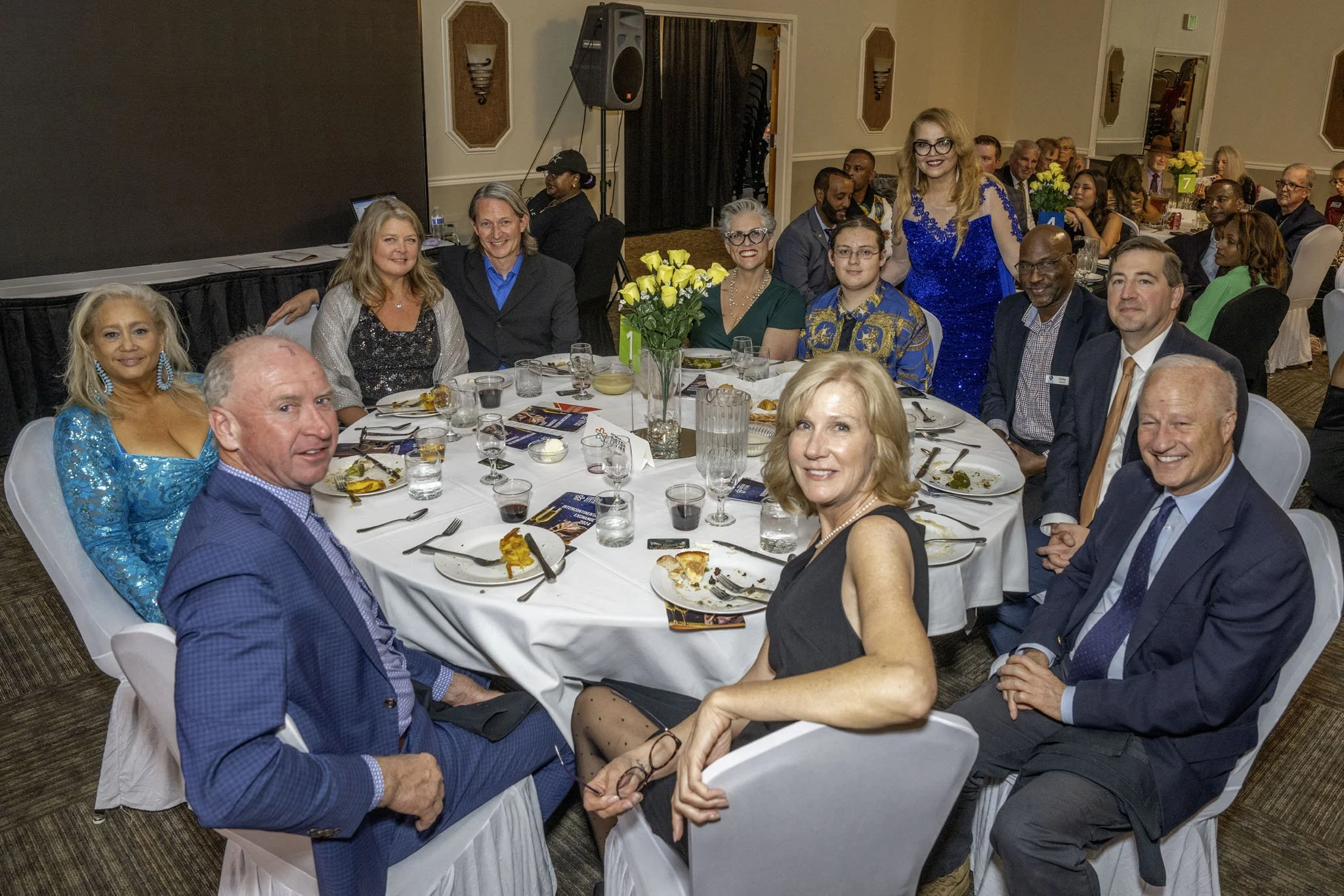 People dressed in formal attire sitting and standing around a banquet table at a formal event or dinner party, with some eating and some posing for the photo, in a decorated banquet hall.