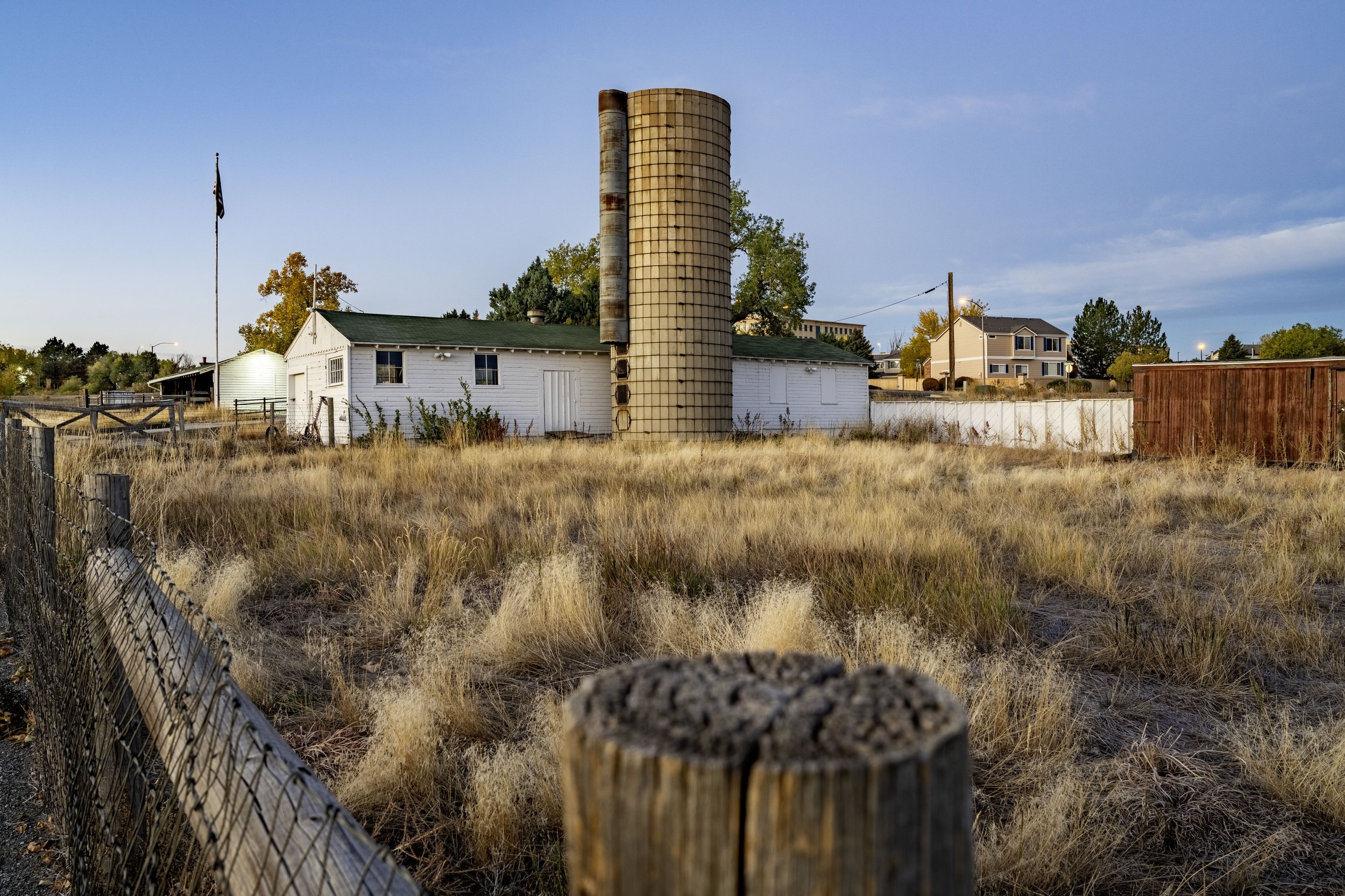 A rural scene with a white house, a rusty silo, and dry grass in the foreground under a partly cloudy sky.