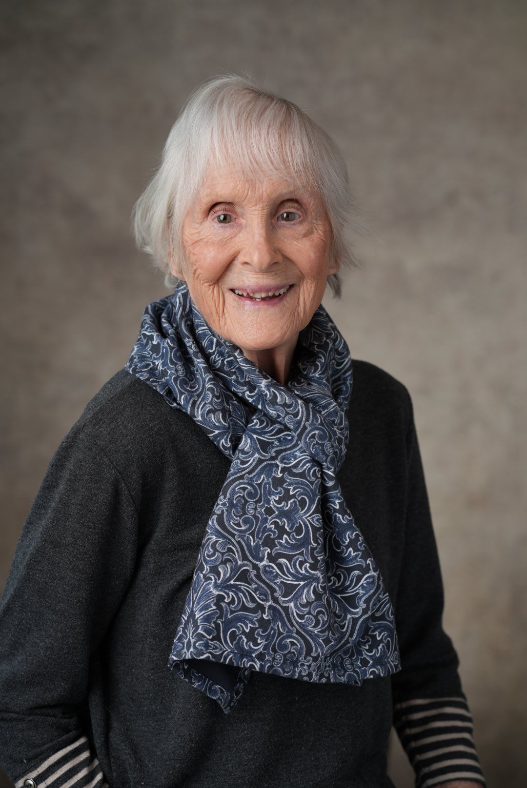 An elderly woman with short, gray hair, wearing a black top and a blue patterned scarf, smiling at the camera.