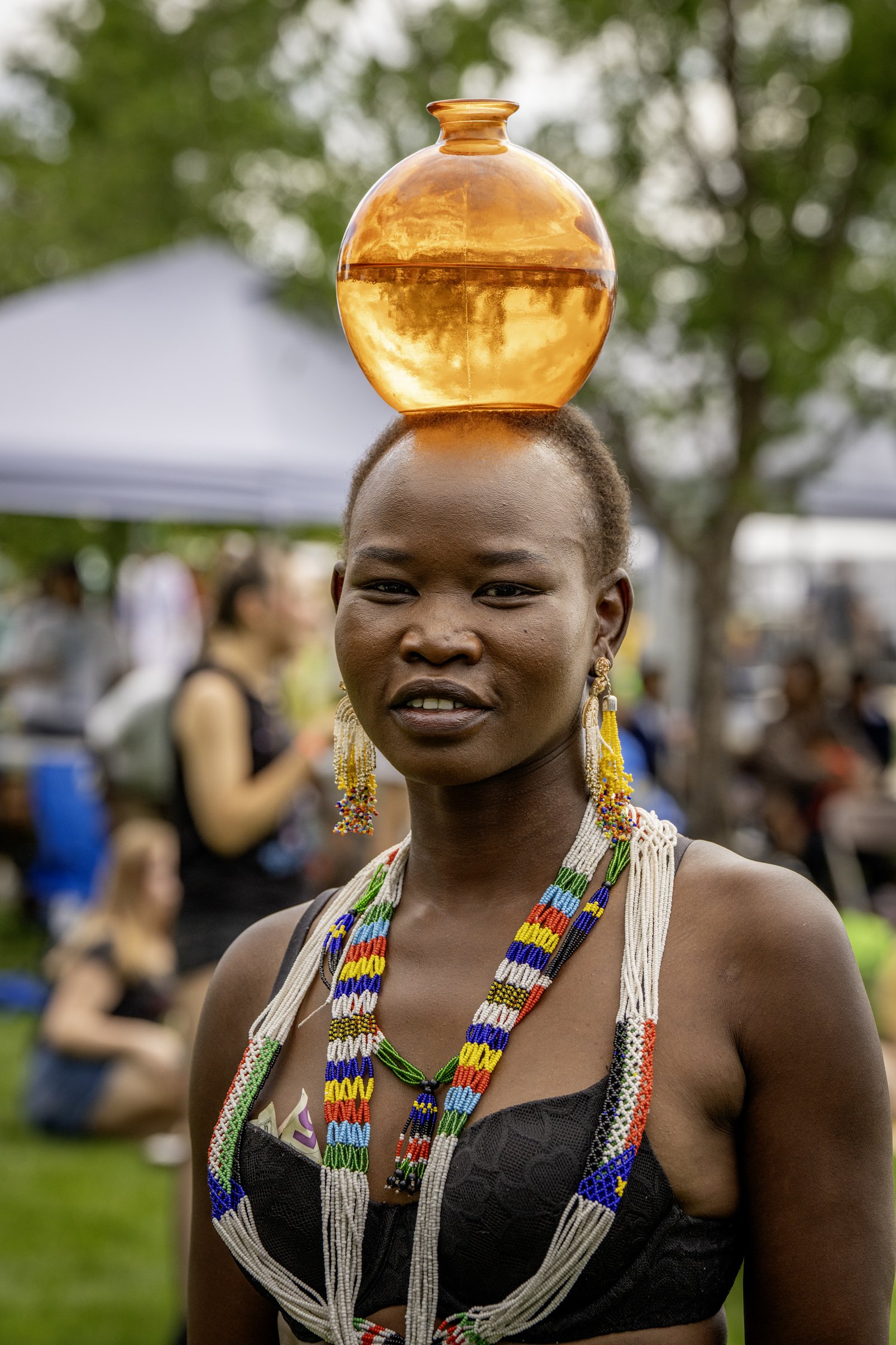 A woman with beaded jewelry and earrings balances a glass bowl filled with water on her head at an outdoor event, with blurred people and tents in the background.