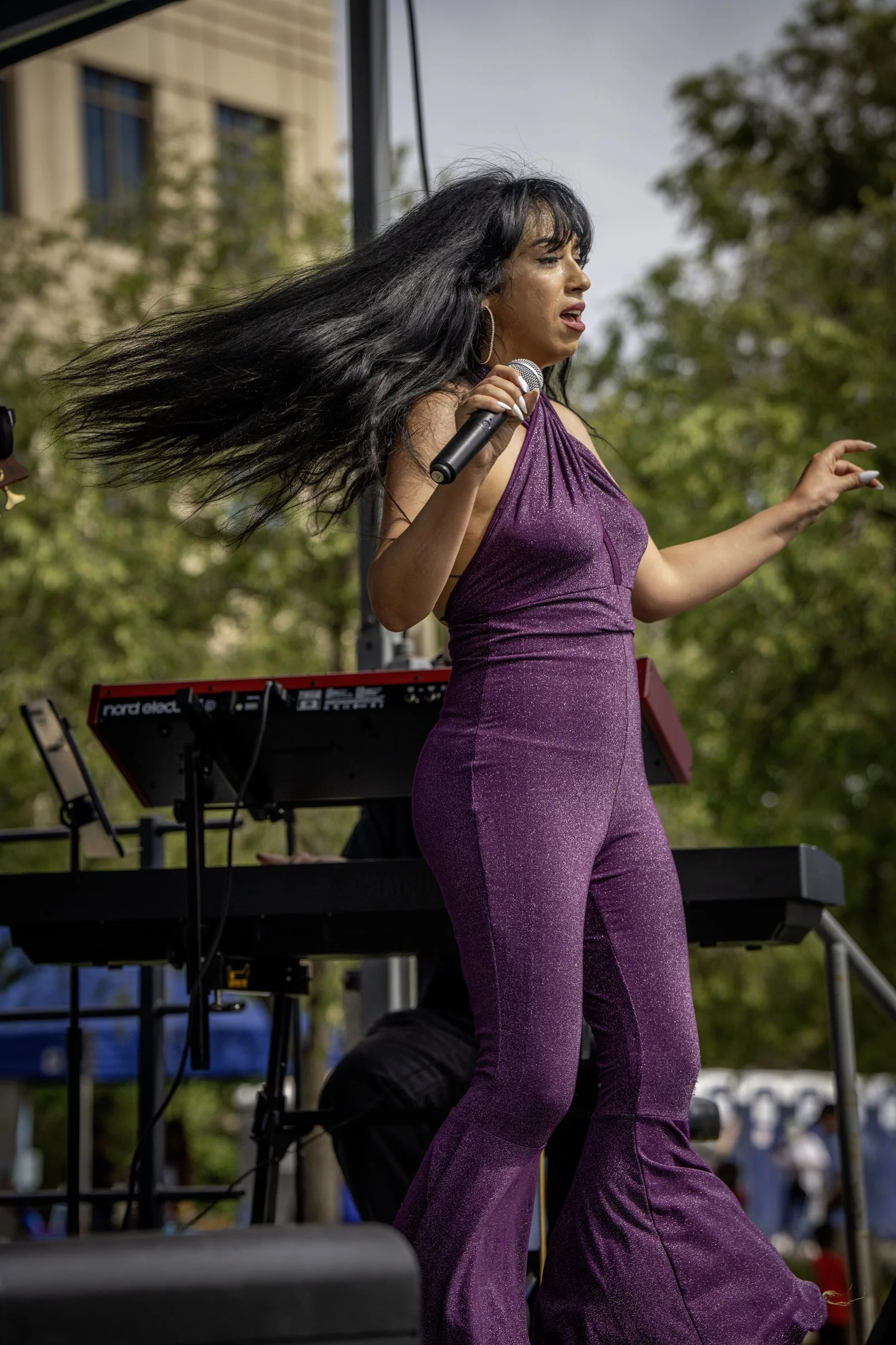 A woman in a purple jumpsuit singing into a microphone on an outdoor stage with a keyboard and green trees in the background.