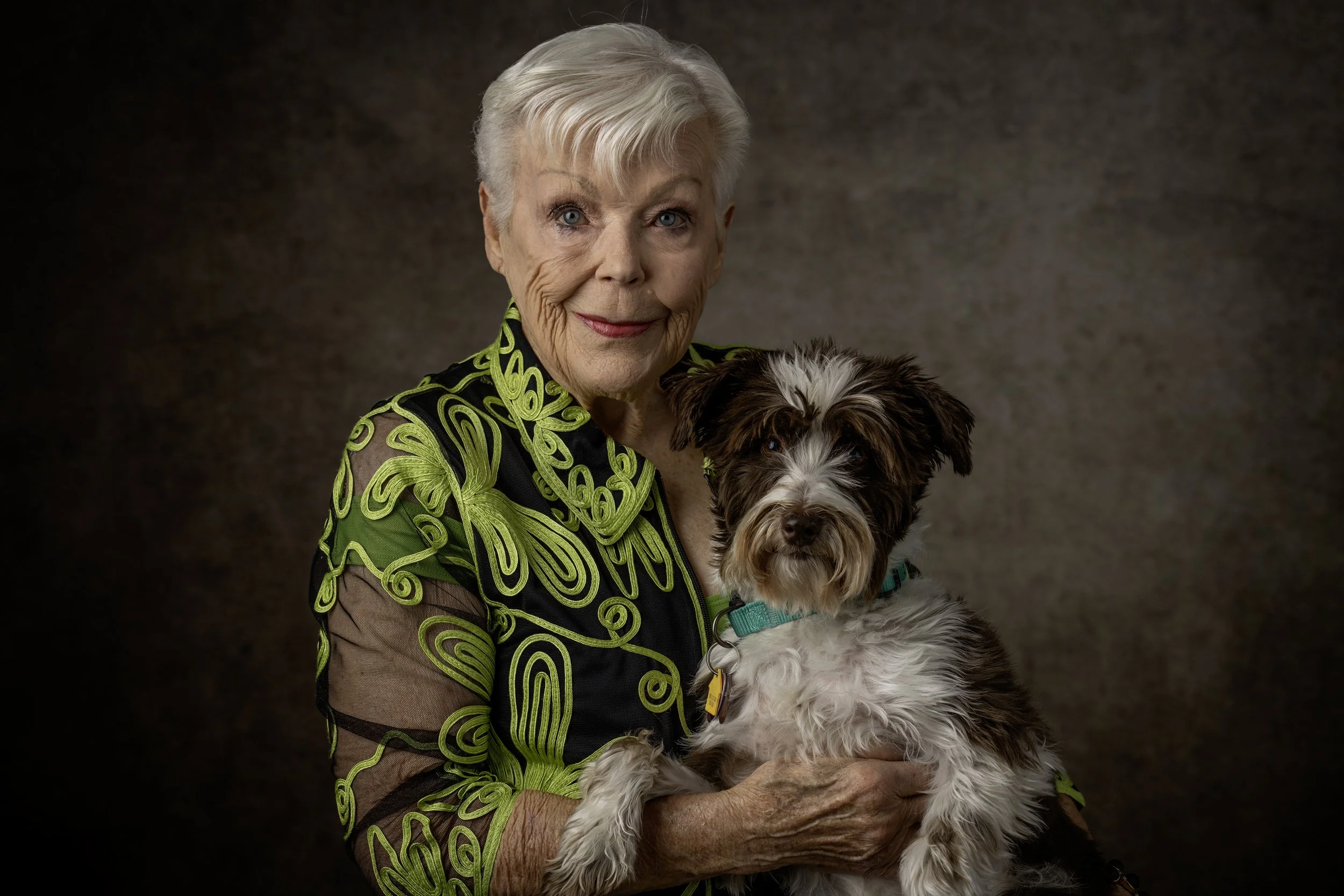 An elderly woman with short gray hair holding a small brown and white dog in her arms. The woman is wearing a black and green patterned blouse, and both are looking at the camera against a dark, textured background.