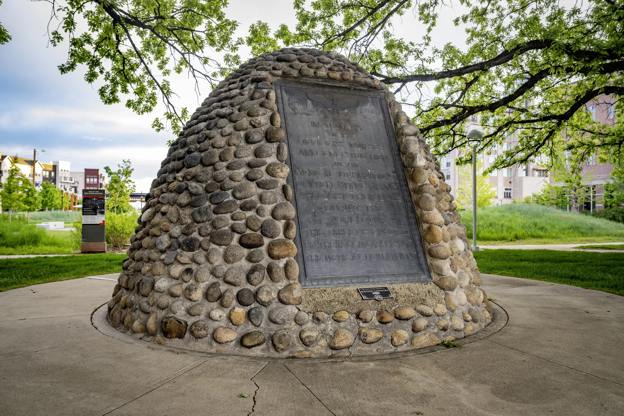 Memorial monument made of rounded stones with metal plaque in an urban park setting, surrounded by greenery and trees.
