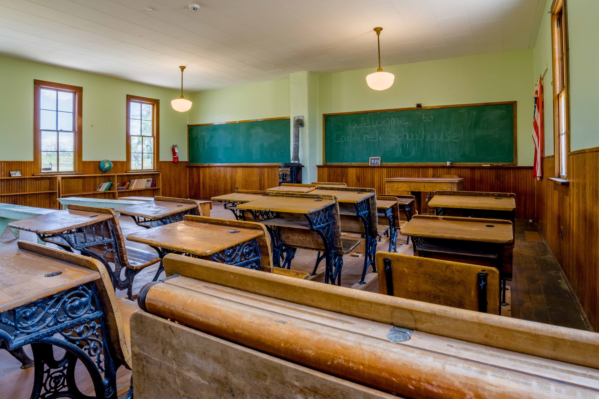 Empty classroom with wooden desks and chairs, green chalkboard, wooden wainscoting, windows with grid panes, a globe, and a flag. Welcome message written on the chalkboard.