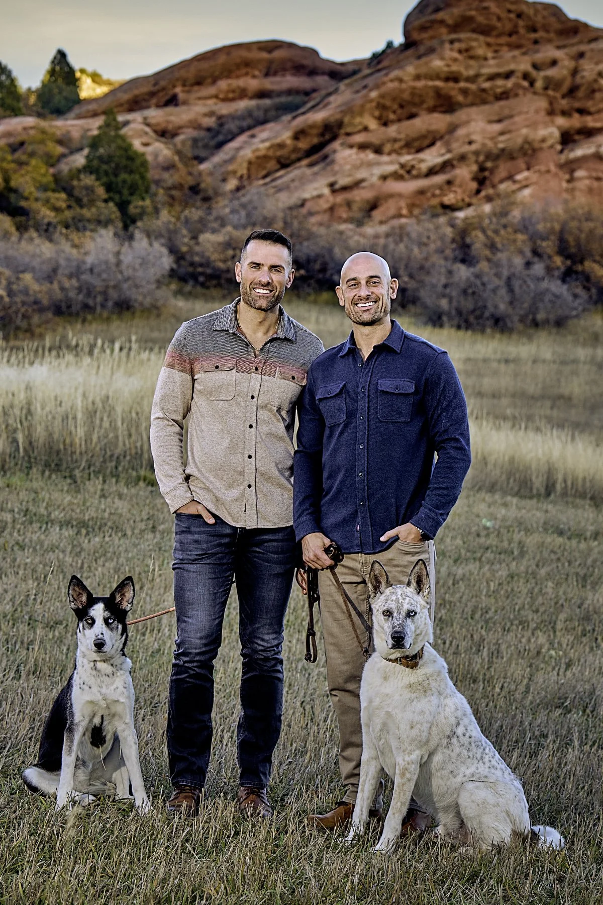 Two men standing outdoors in a grassy field with two dogs, one sitting and one standing, with red rock formations and trees in the background during sunset.