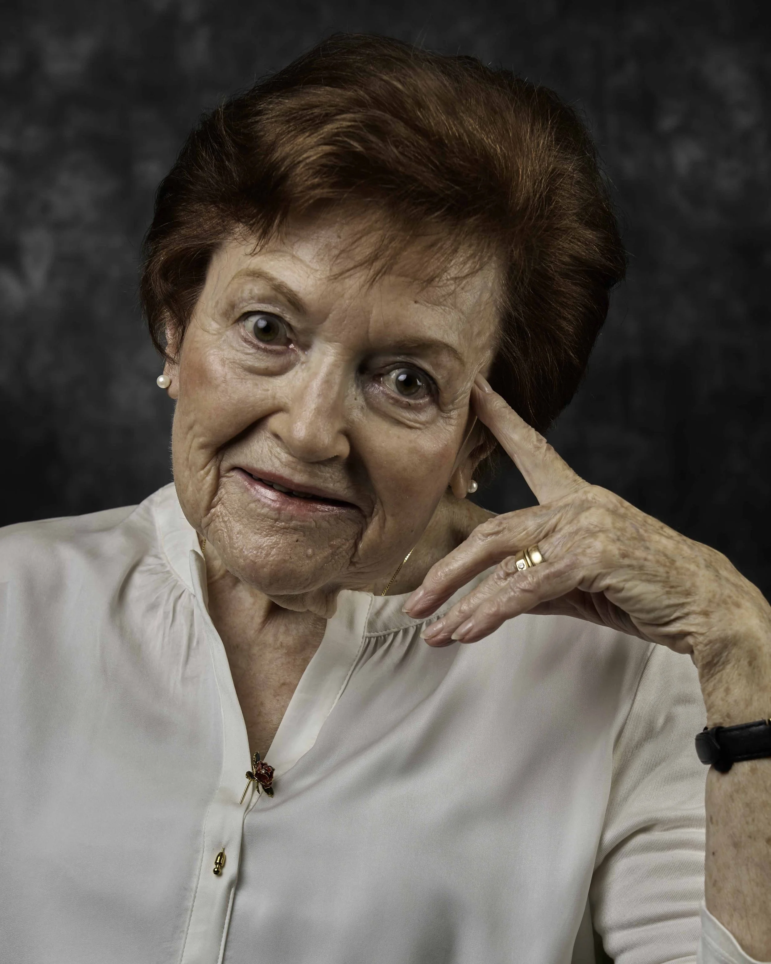 An elderly woman with short, auburn hair and light skin, gently touching her temple with her right hand, looking directly at the camera with a slight smile, wearing a white blouse, pearl earrings, a gold ring, and a black wristwatch, against a dark, 