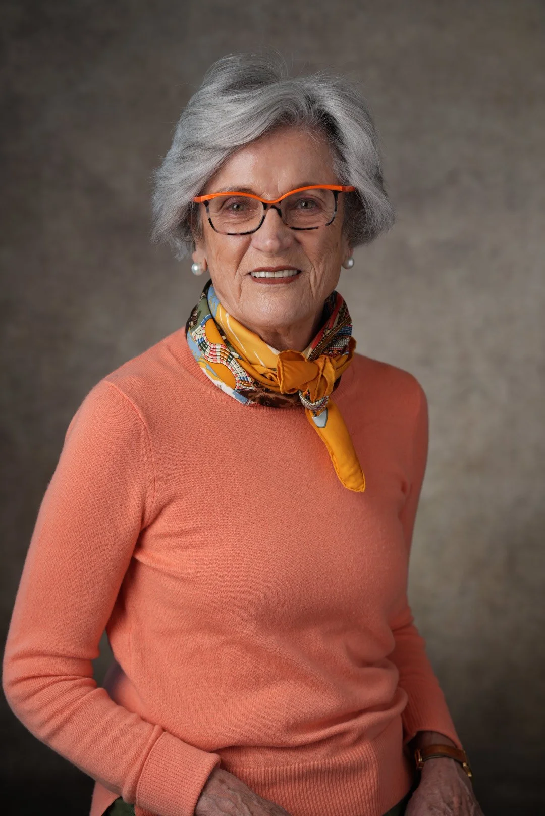 A portrait of an elderly woman with gray hair, wearing glasses with orange accents, a peach sweater, a colorful scarf around her neck, pearl earrings, and a watch, standing against a plain background.
