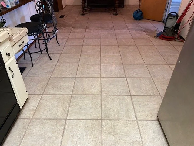 Kitchen floor with beige tiles and black grout lines, with a section of counter and chairs on the left, and cleaning equipment on the right.