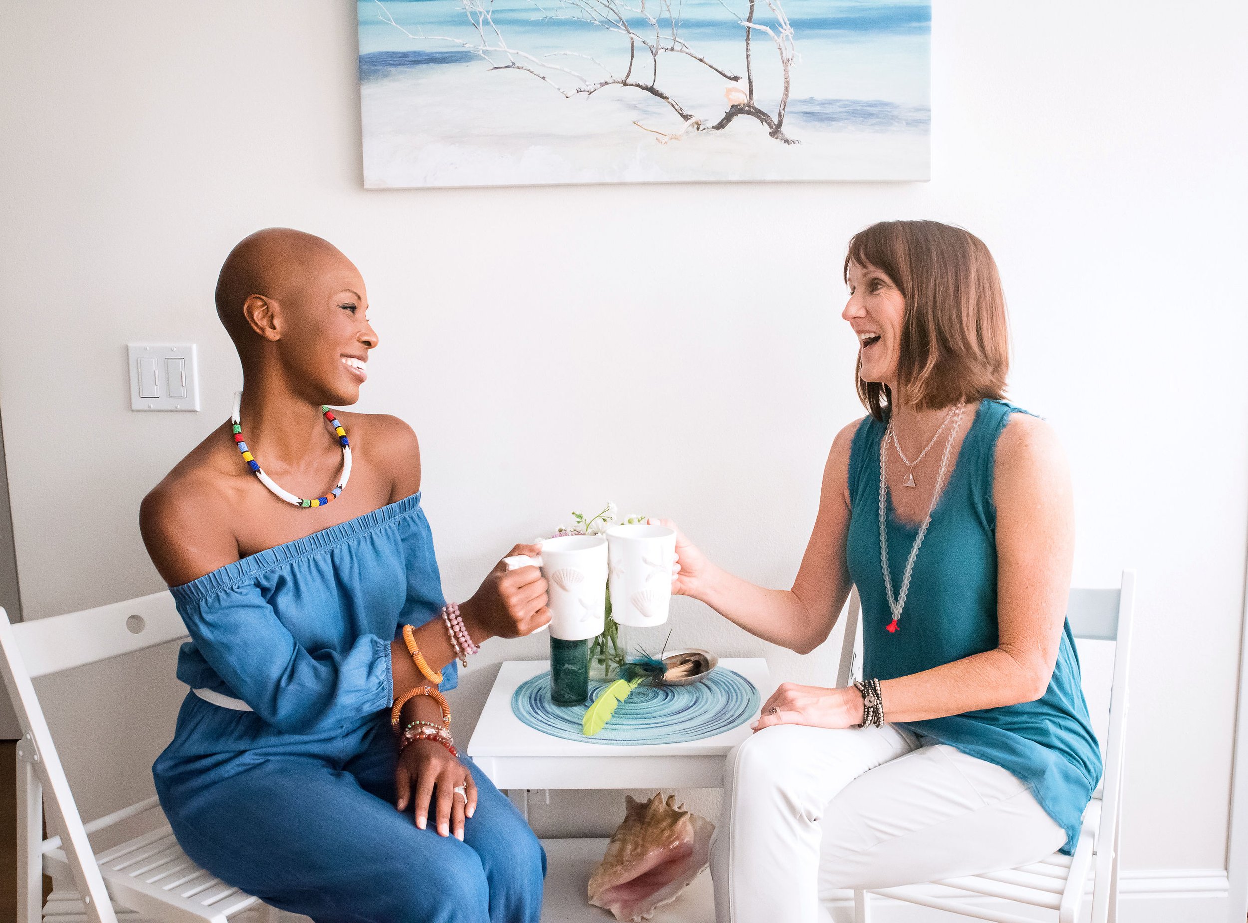 Onyi Love and her colleague sitting at a white table, smiling and holding white mugs, toasting each other, with a painting of a beach scene on the wall behind them.