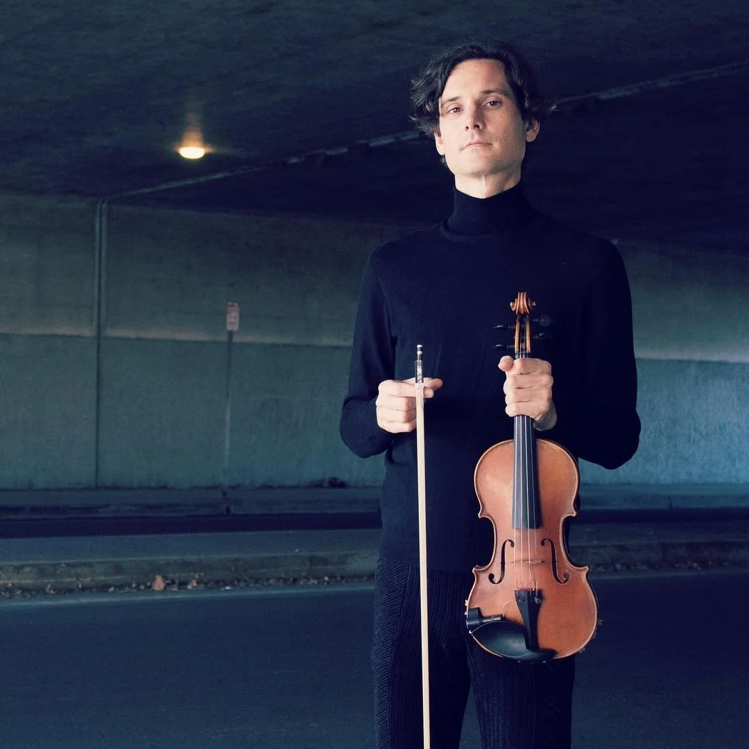 Violinist Greggory Allison wearing a black turtleneck holding a violin and a bow, standing under an overpass.