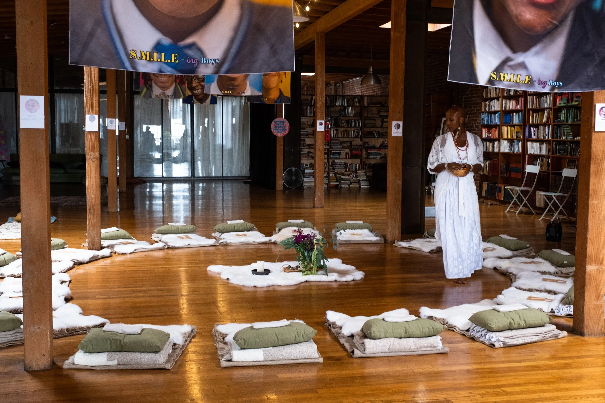 Indoor space set up with pillows and blankets arranged in a circle on a wooden floor, with a Onyi dressed in white standing near the arrangement, bookshelves in the background, and banners hanging from the ceiling.
