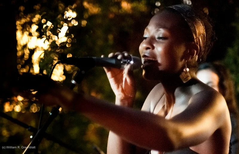 Onyi Love singing into a microphone outdoors at night, with a blurred background of trees and warm lighting.