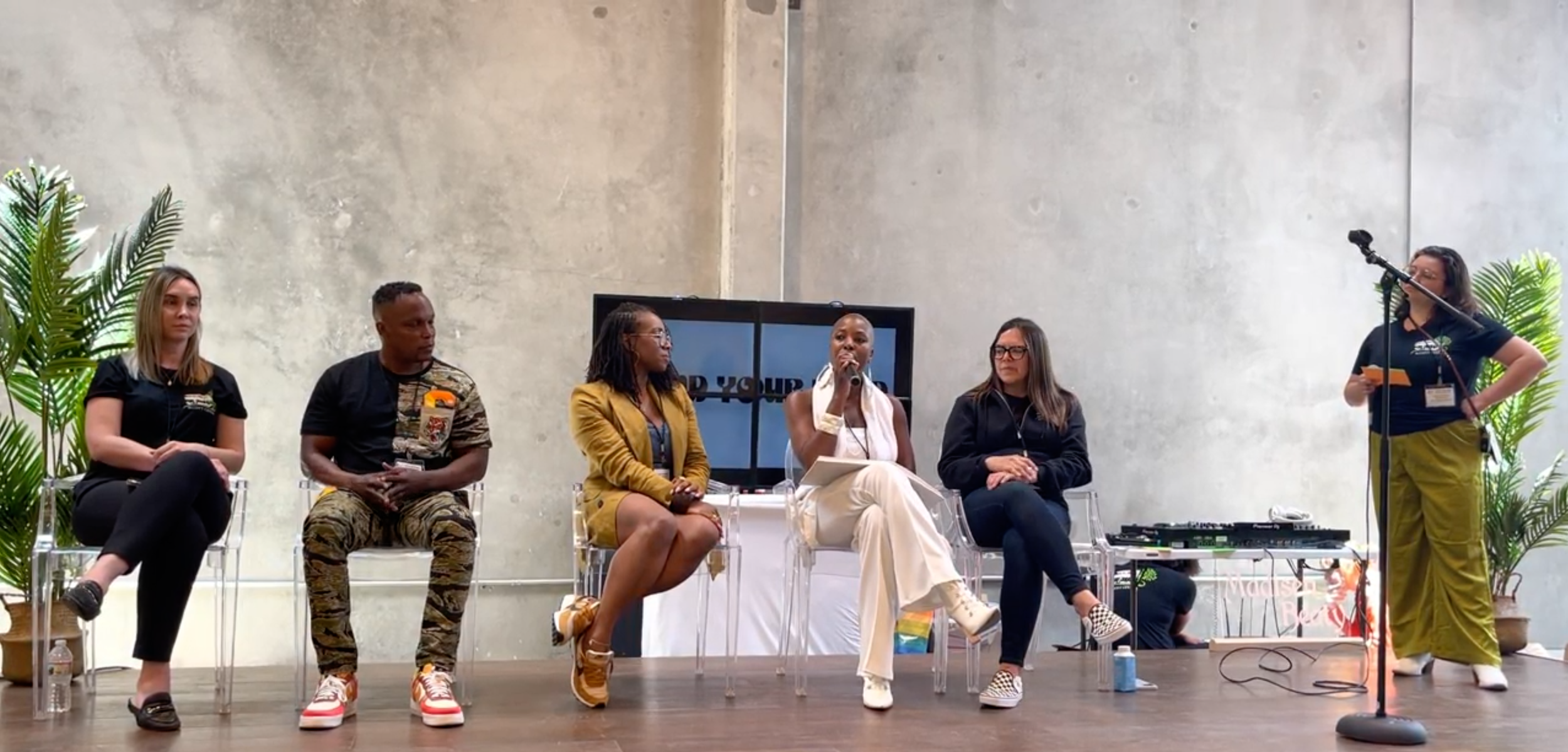 A panel discussion with five people, including Onyi Love, healer and consultant seated on stage. One person is standing at a microphone, against a plain wall with two large potted plants.