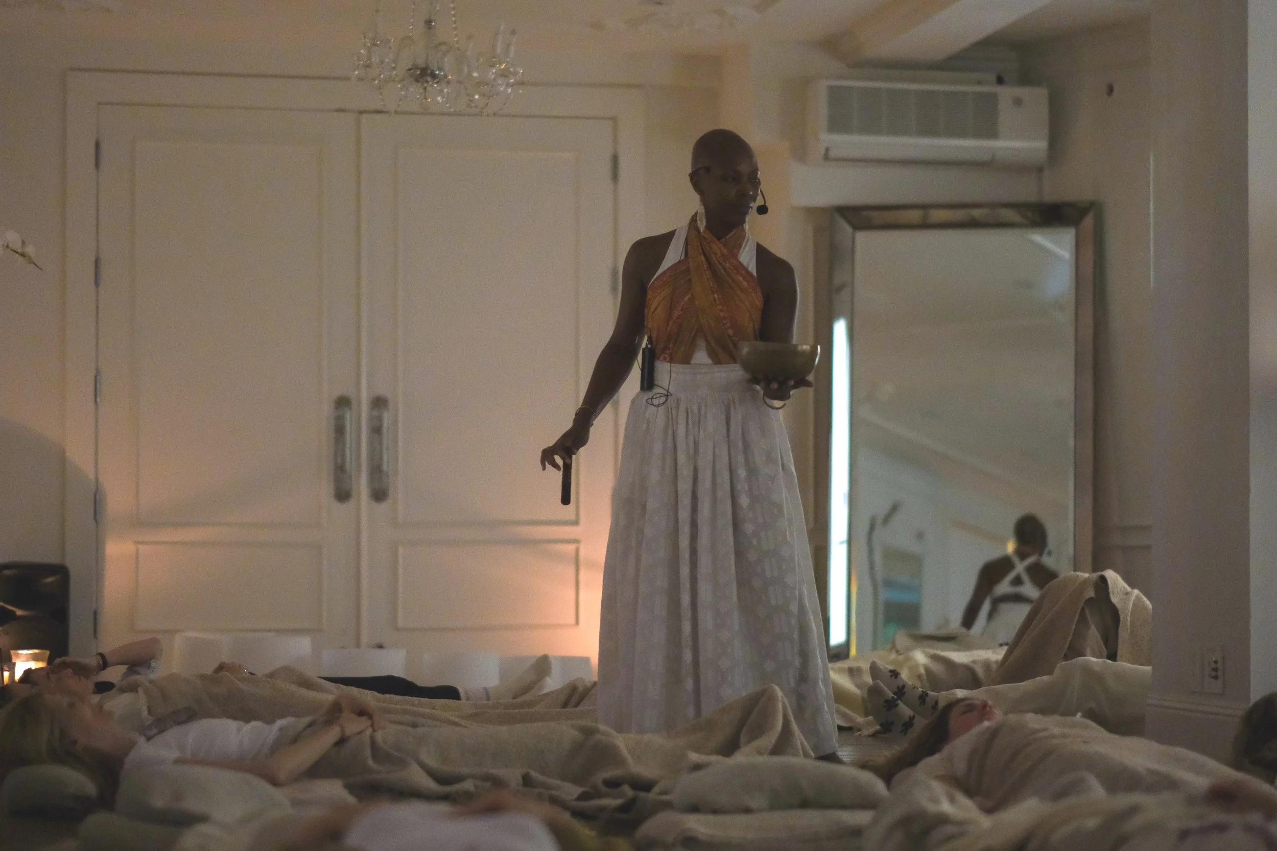 Healer and priestess Onyi Love standing in a room holding a bowl and a microphone, with several people lying down covered with blankets on the floor around her.