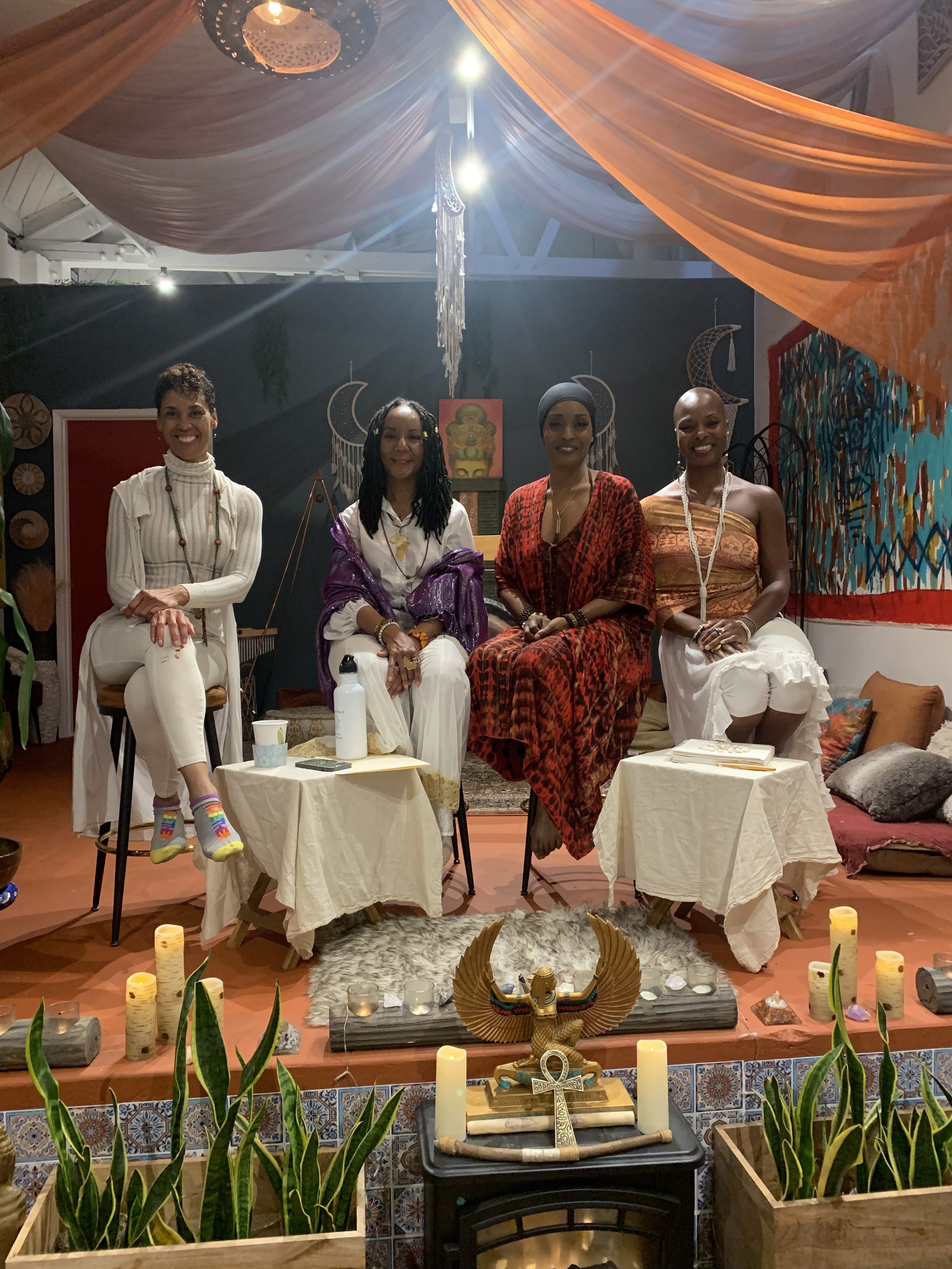 Onyi Love and 3 colleagues sit on chairs arranged in a semi-circle in a room decorated with colorful fabrics, art, candles, and plants. They are panelists for a talk on Spiritual Abuse.