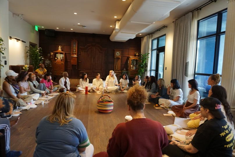 Onyi Love and a group of diverse women sitting in a circle on the floor during a workshop or discussion in a spacious, well-lit room with wooden floors and large windows.