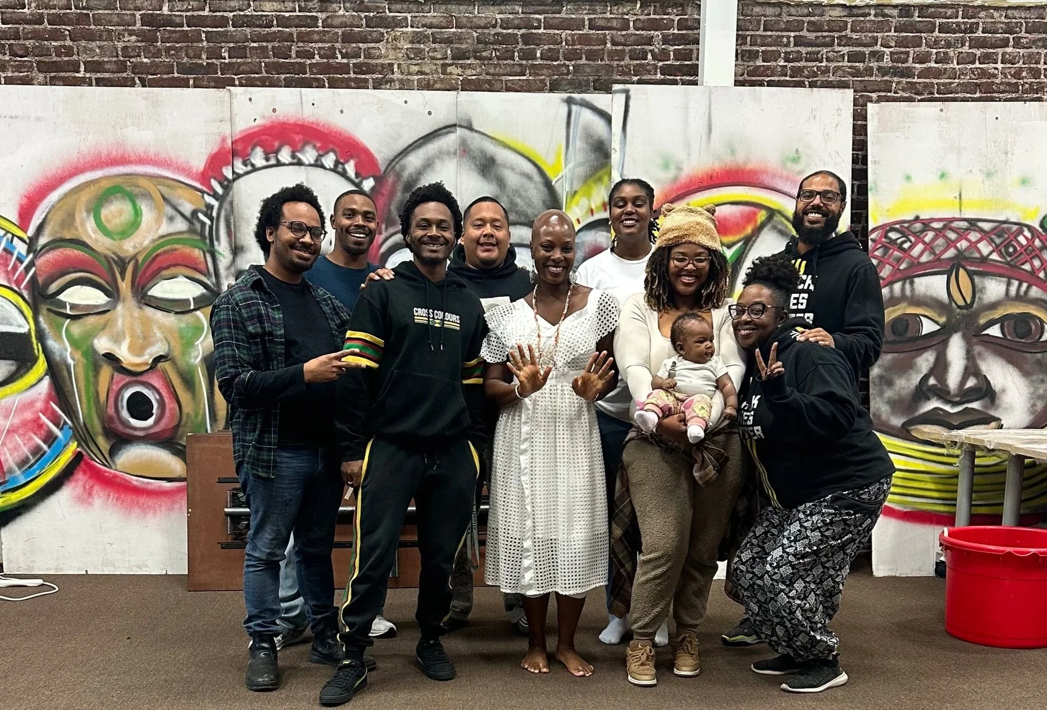 9 adults, including a woman and her infant, and healing Onyi pose in front of a brick wall. Leaning against the wall are wooden boards with indigenous African faces or tribal masks painted on them.