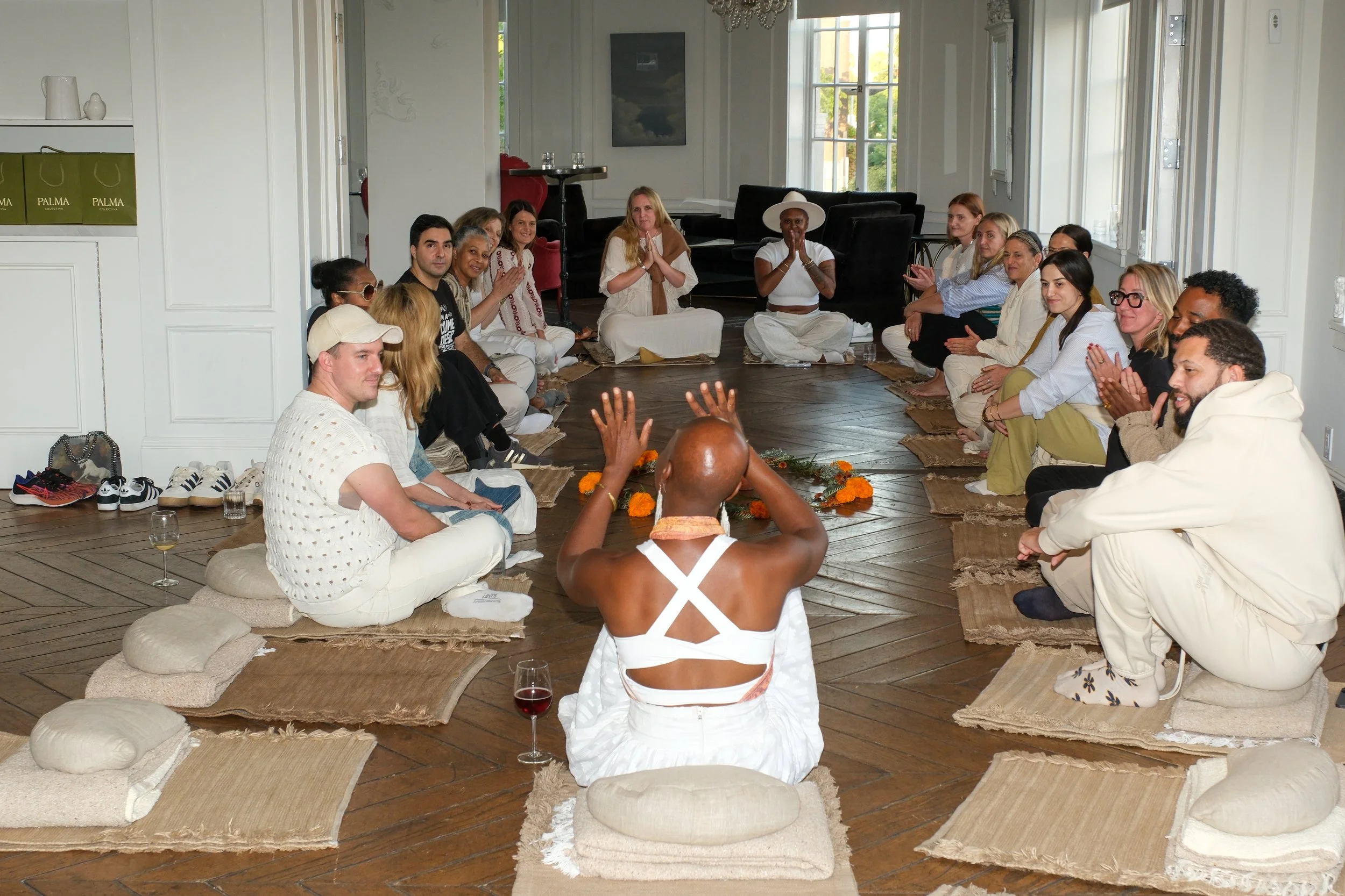 Onyi sitting with a group in circle on cushions and mats, participating in a yoga or meditation session in a bright room with large windows and wooden floor.