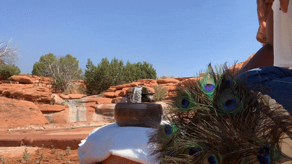 Onyi Love playing a metal singing bowl near a waterfall at the Sedona Mago Retreat Center. A peacock displaying its feathers is beside her. The surroundings are a desert landscape with rocks, shrubs, and a clear blue sky.