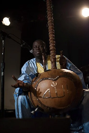 Musician Amadou Fall in traditional clothing plays a kora, a West African string instrument, on stage with lighting and a microphone setup.