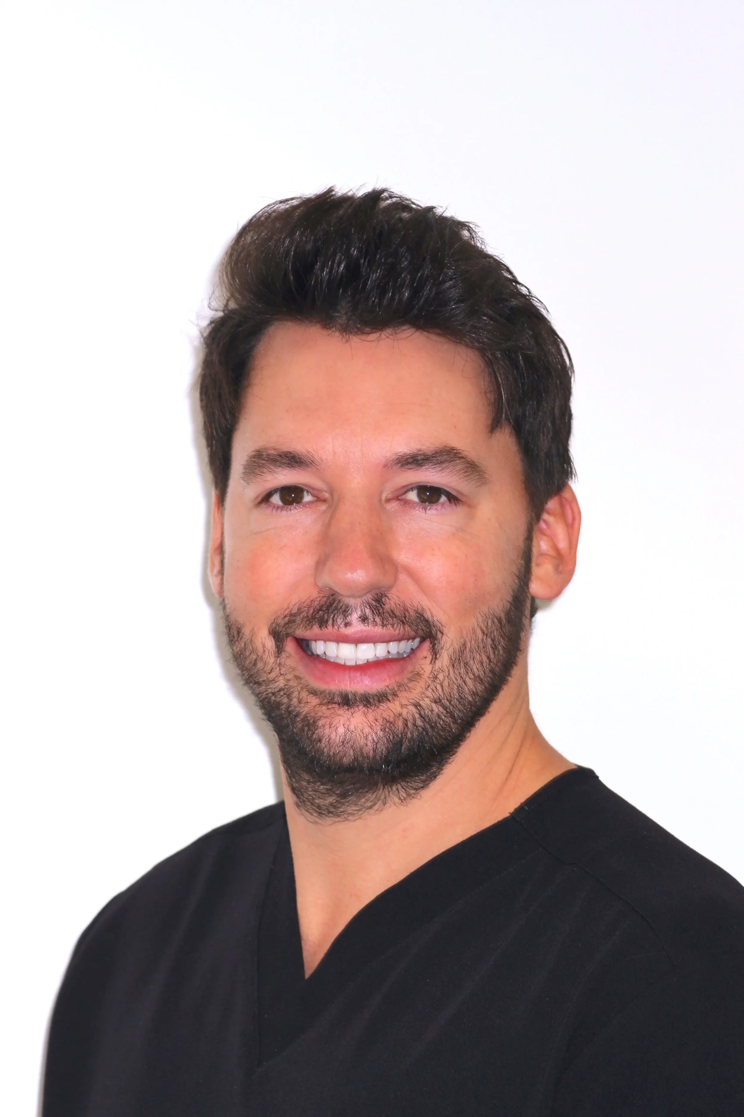 Close-up of a smiling man with dark, styled hair, a beard, wearing a black V-neck shirt, against a plain white background.