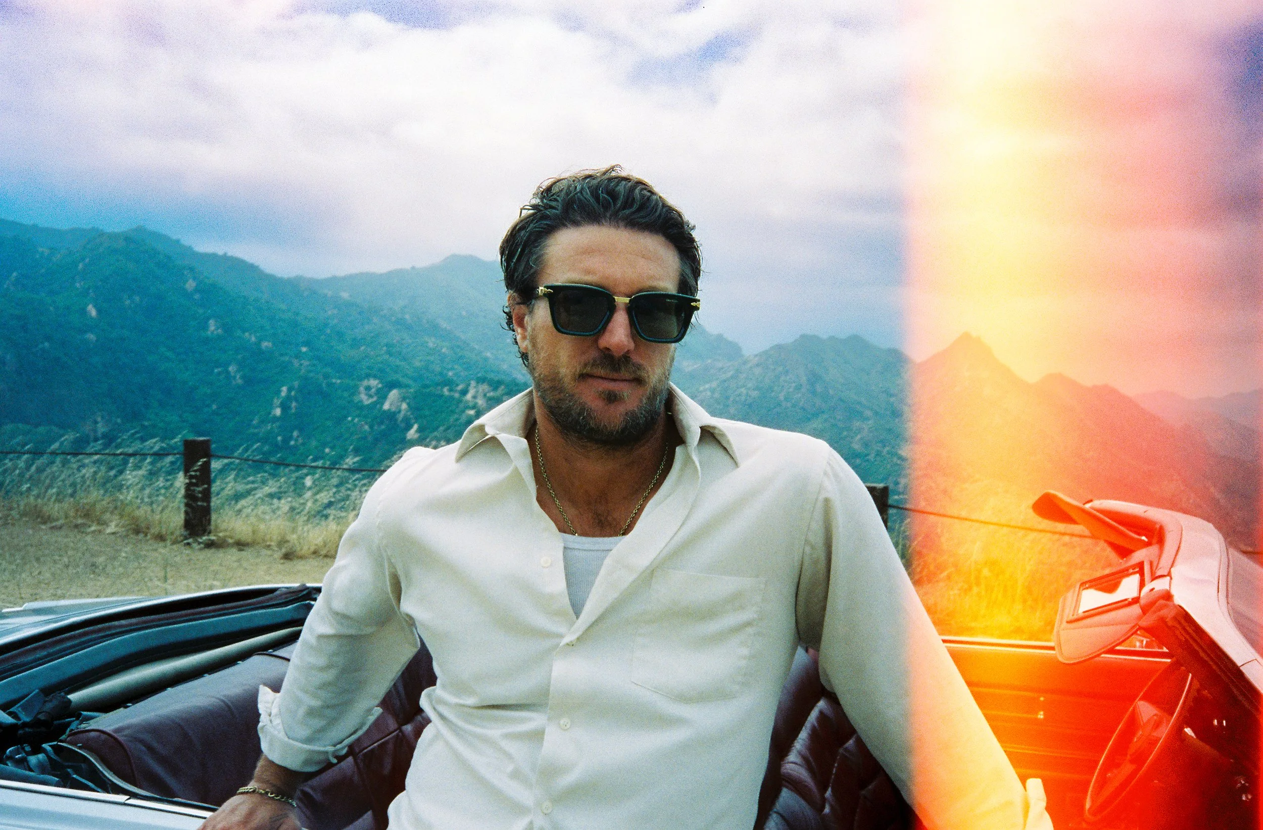 A man with dark hair and beard wearing sunglasses and a white shirt, sitting in a convertible car with mountains and cloudy sky in the background.