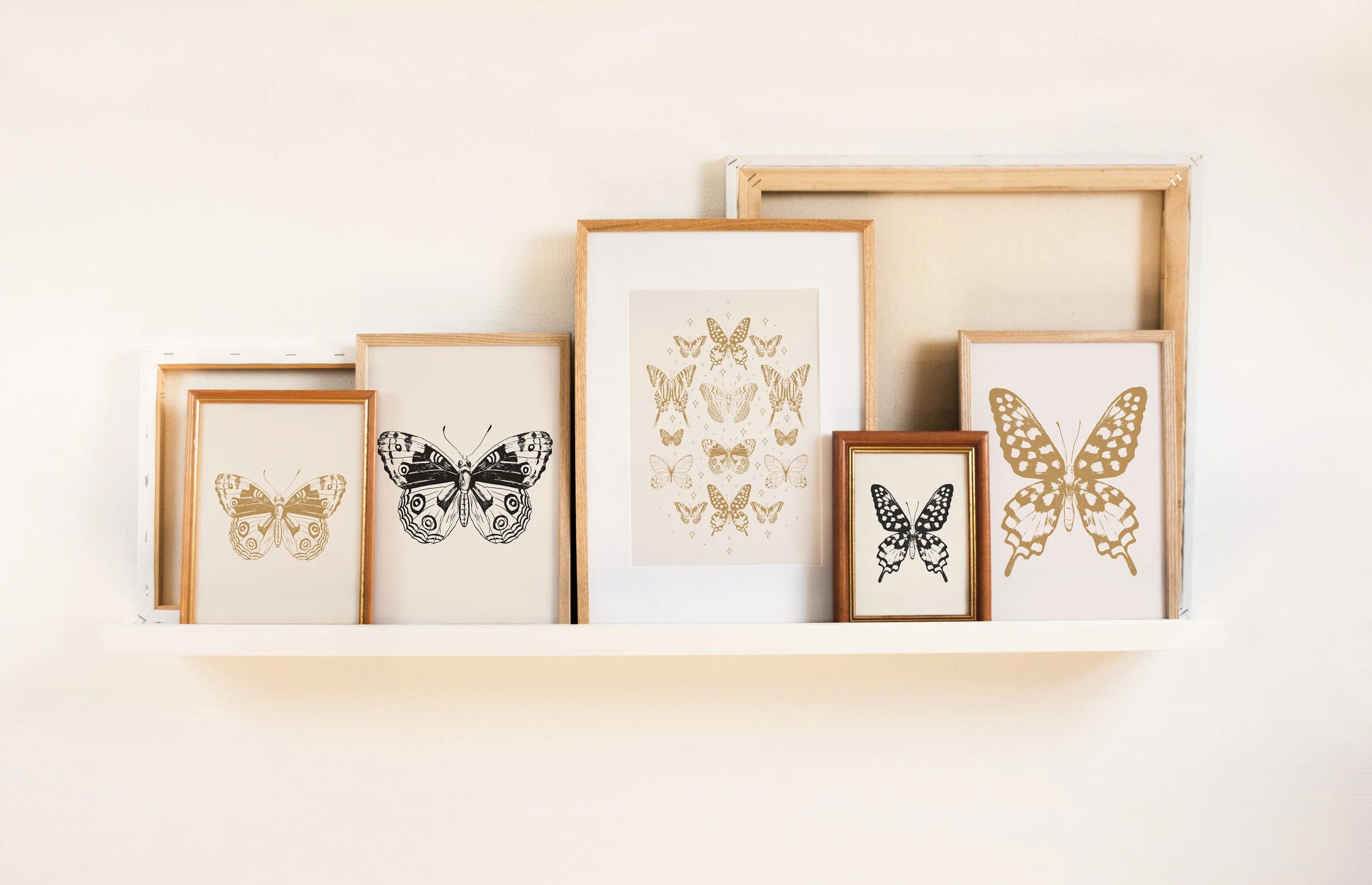 A collection of framed butterfly artwork arranged on a white floating shelf against a white wall. The artwork features black and gold butterfly designs of various sizes and styles.