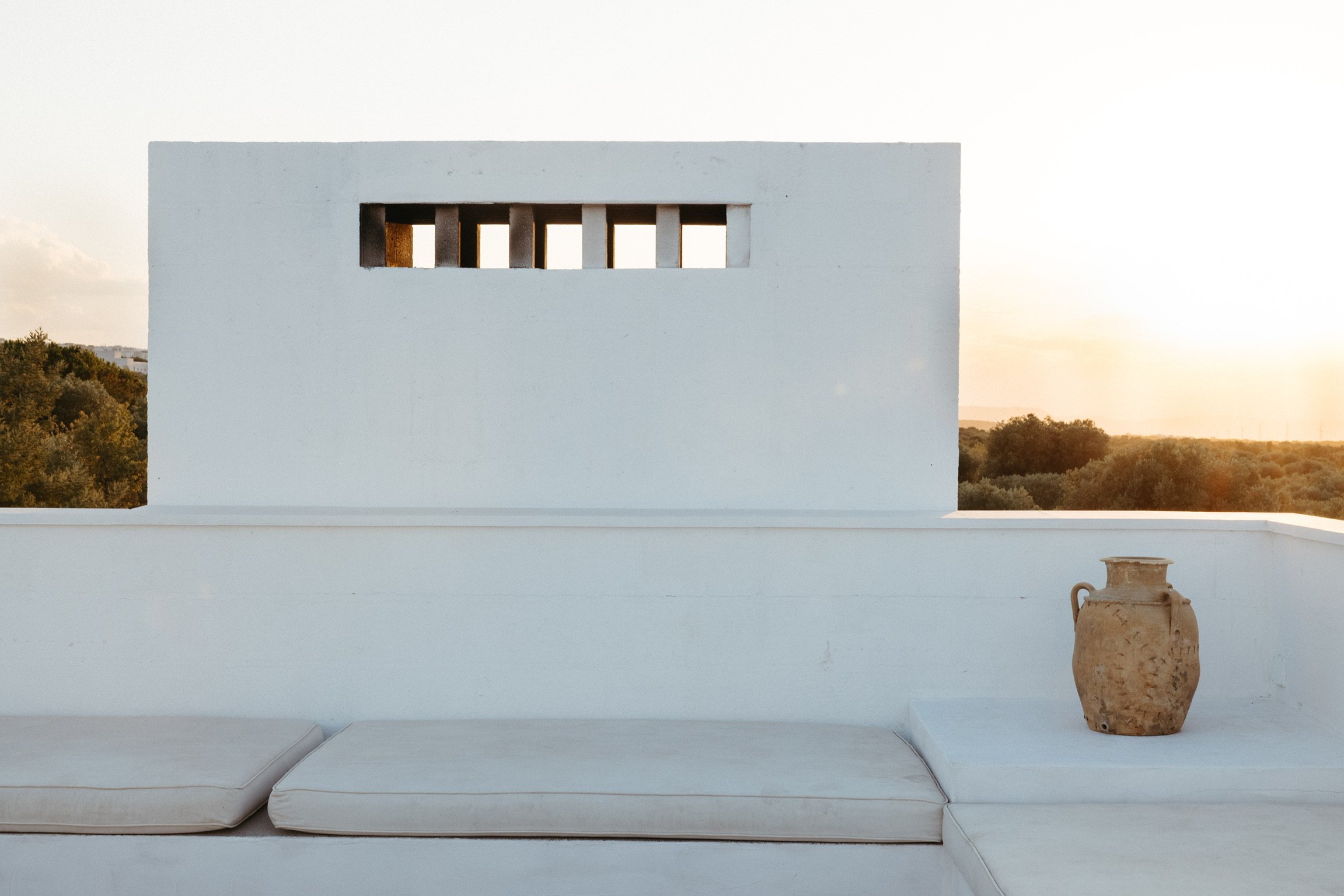 Minimalist outdoor seating area with white cushions, a white wall with vertical rectangular openings, an ancient-looking clay pot, and a view of trees at sunset.