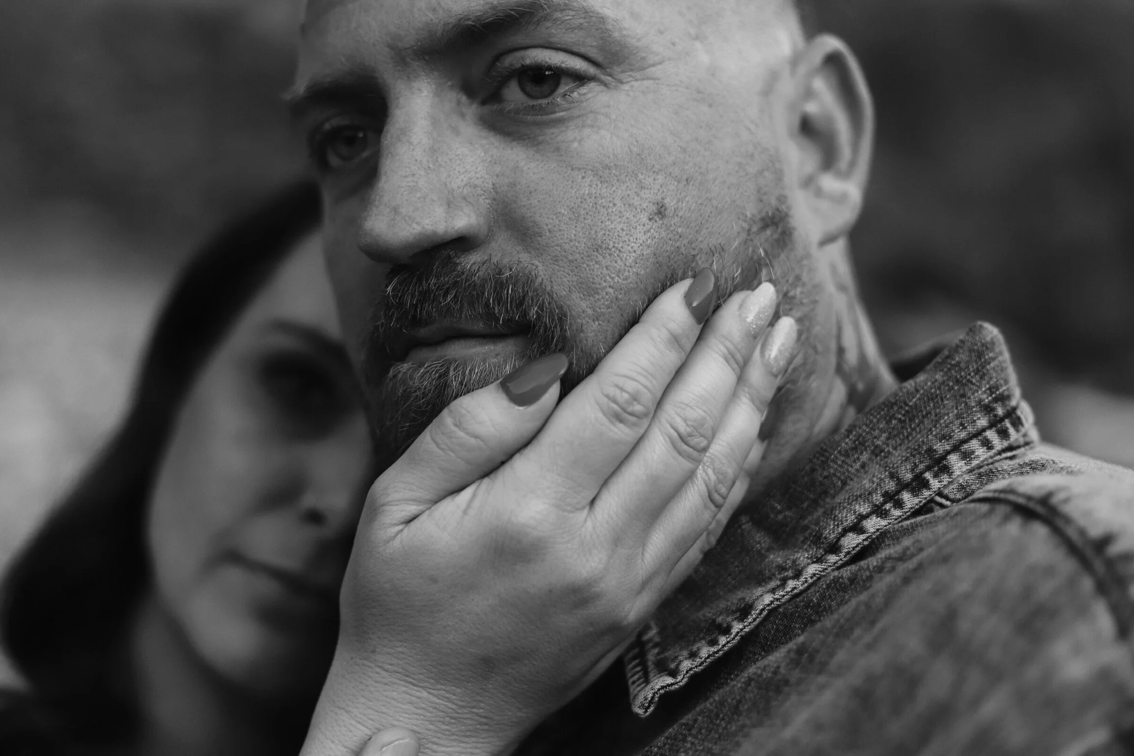 Close-up of a man's face with a contemplative expression, being gently touched by a woman's hand with painted nails, in black and white.