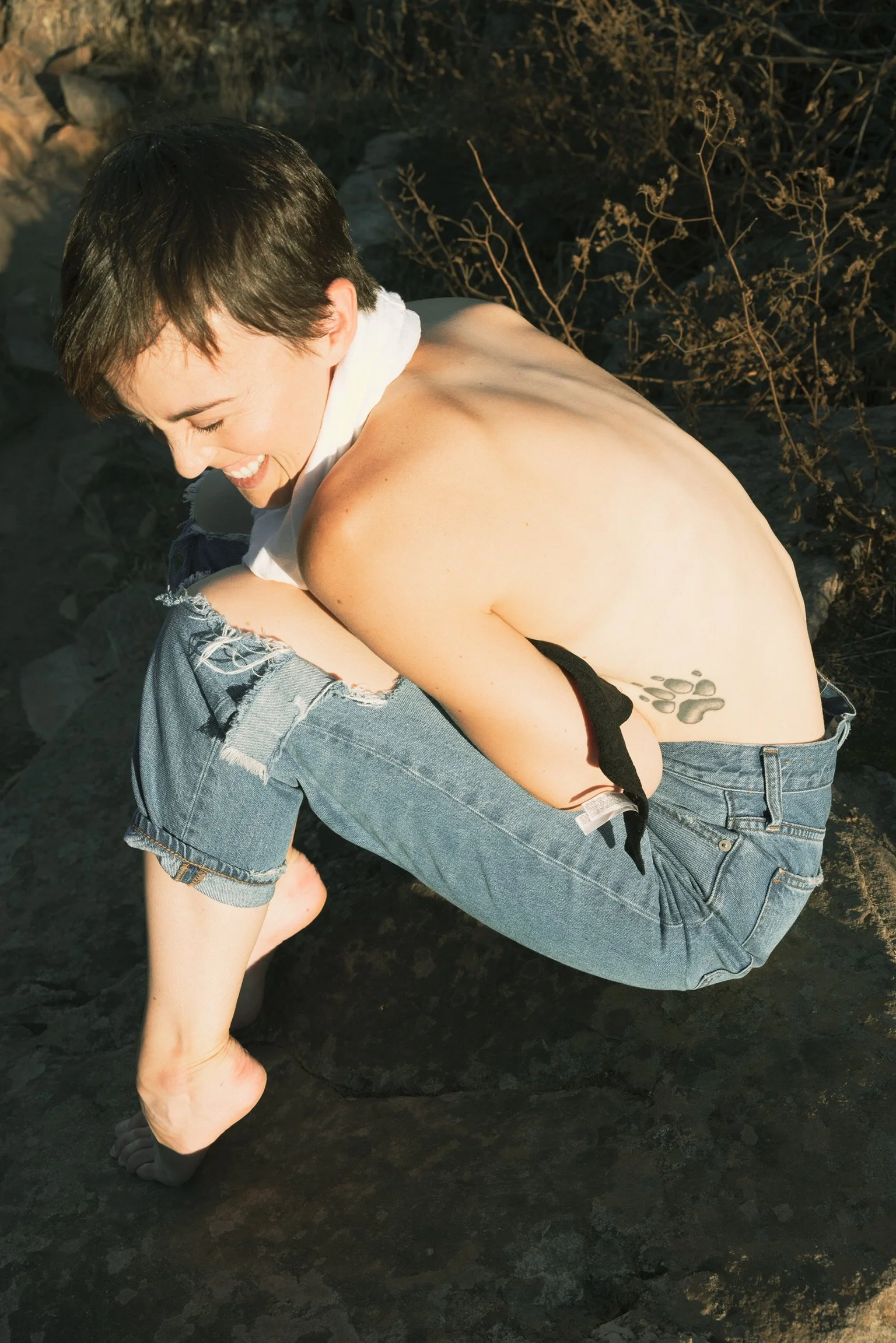 Person sitting outdoors on a rock, wearing ripped jeans, with a tattoo visible on their side.