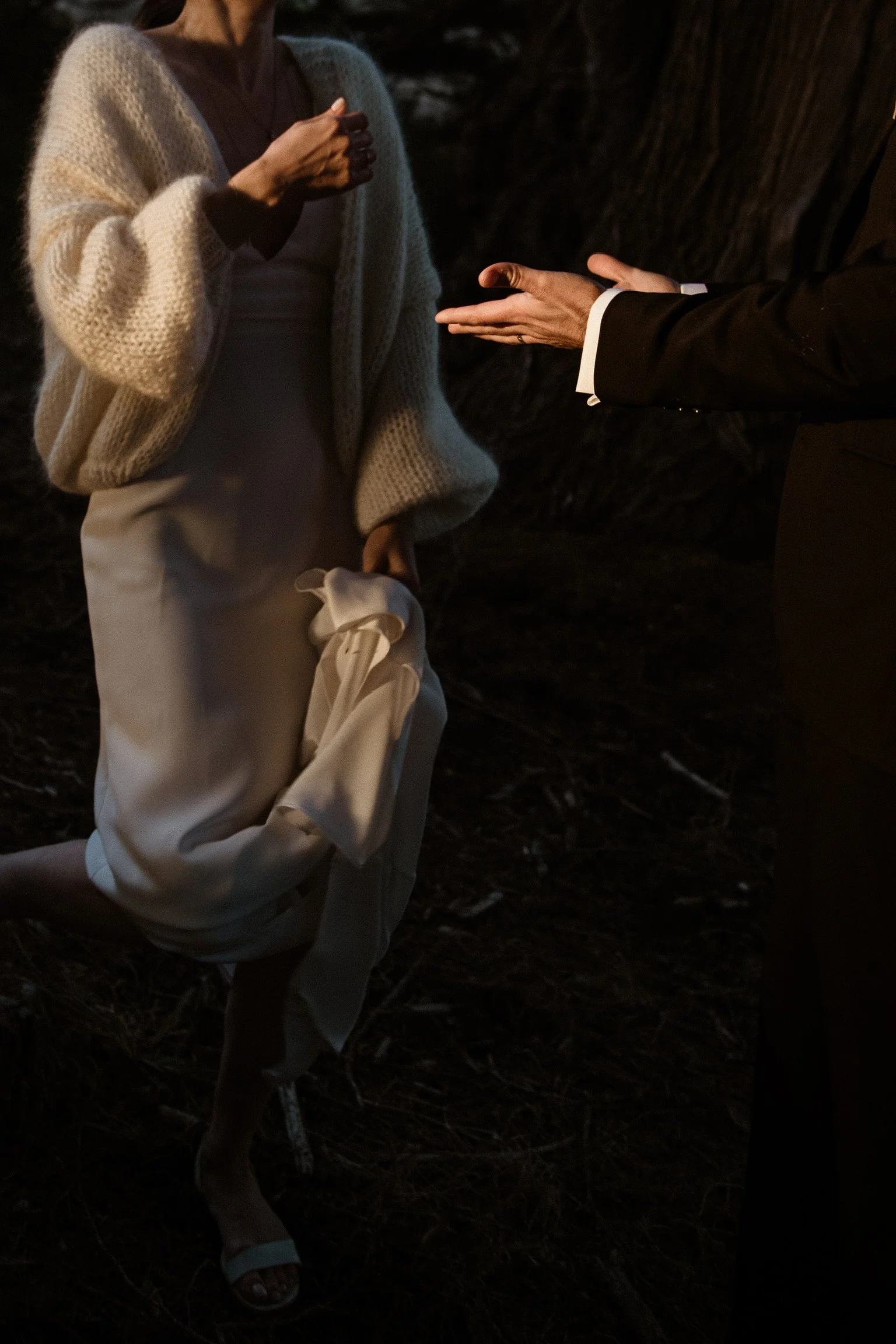 Two people in formal attire, one wearing a knitted sweater and holding a cloth, the other in a suit extending hands, captured in a dimly lit outdoor setting.
