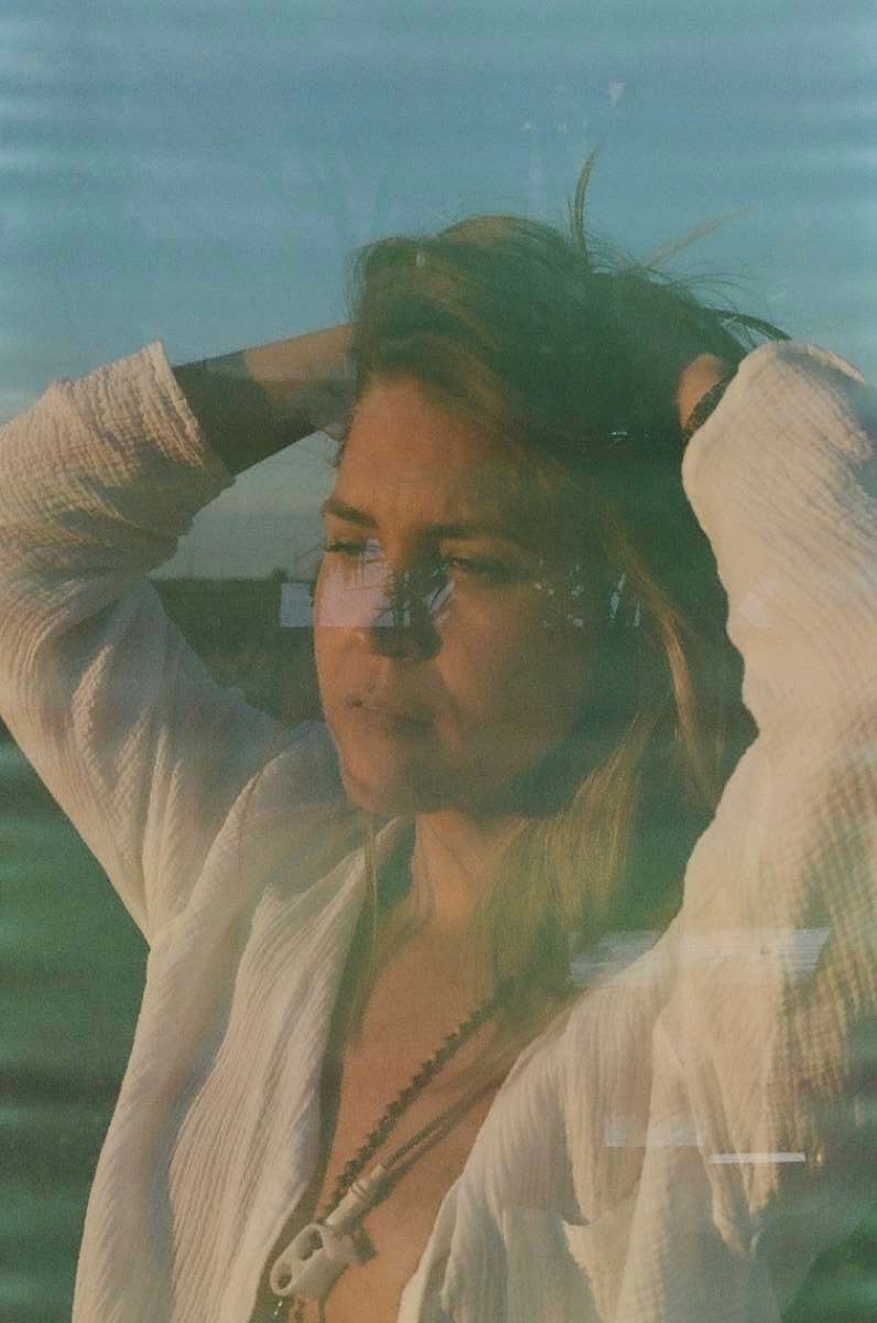 Reflective portrait of a person with hands on head, wearing a white shirt and necklace, seen through glass with a blurred outdoor background.