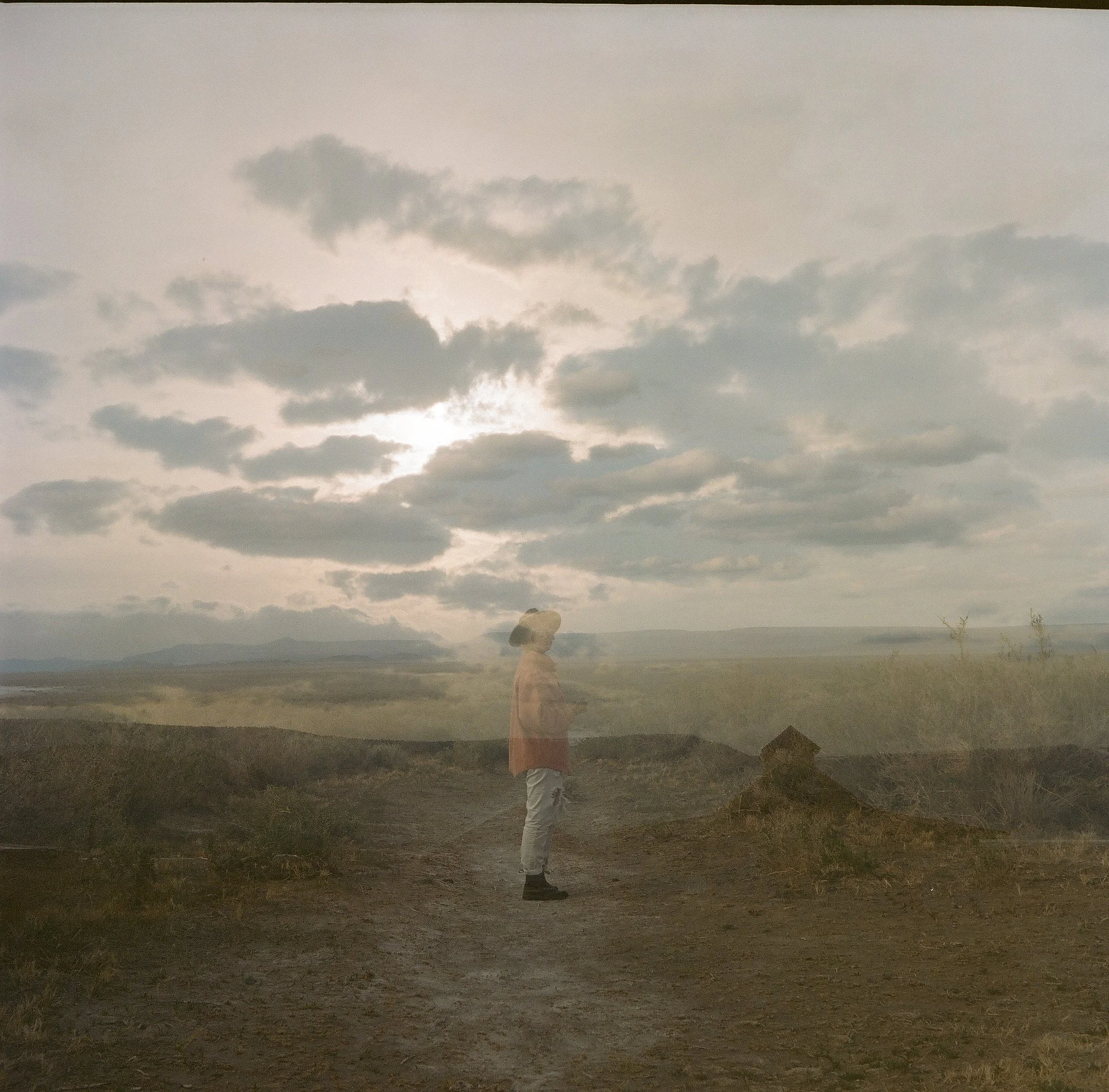 A double exposure image of a person standing on a dirt path, surrounded by an expansive landscape with tall grasses and overcast sky. The image creates a ghostly effect against the cloudy backdrop.