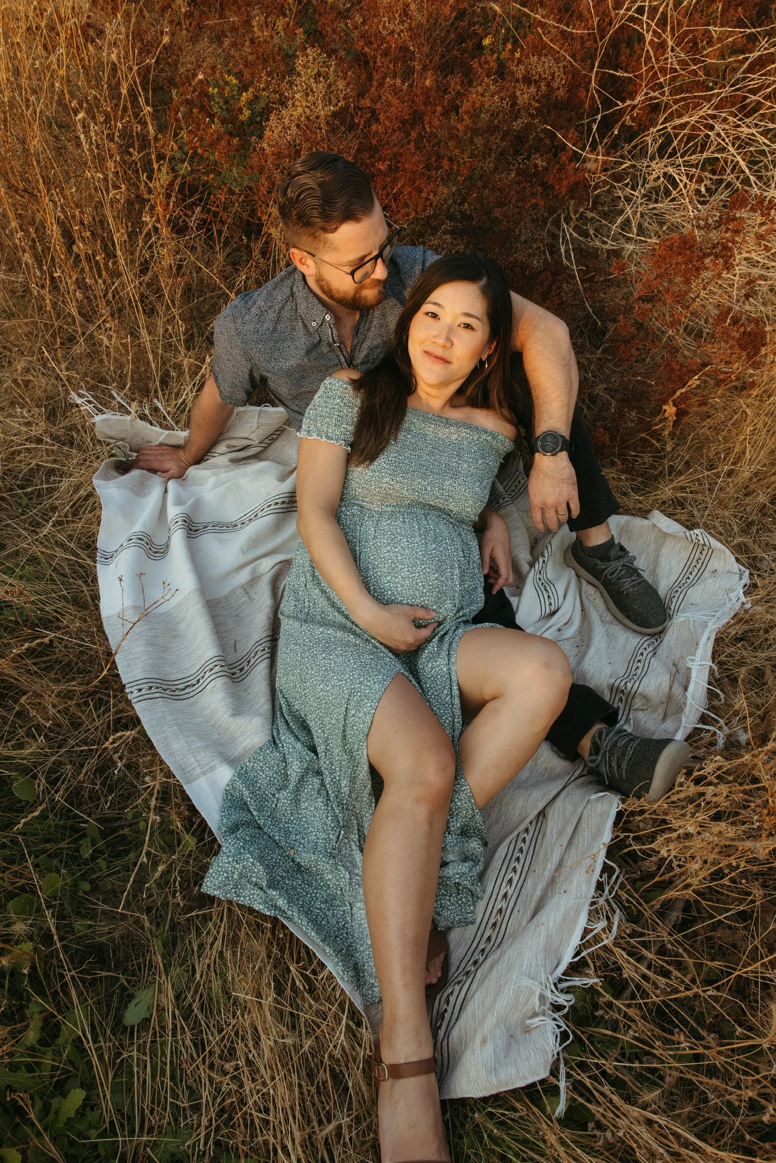 Expectant couple sitting on a blanket in a field, woman in a green dress holding her pregnant belly while leaning against a man embracing her.