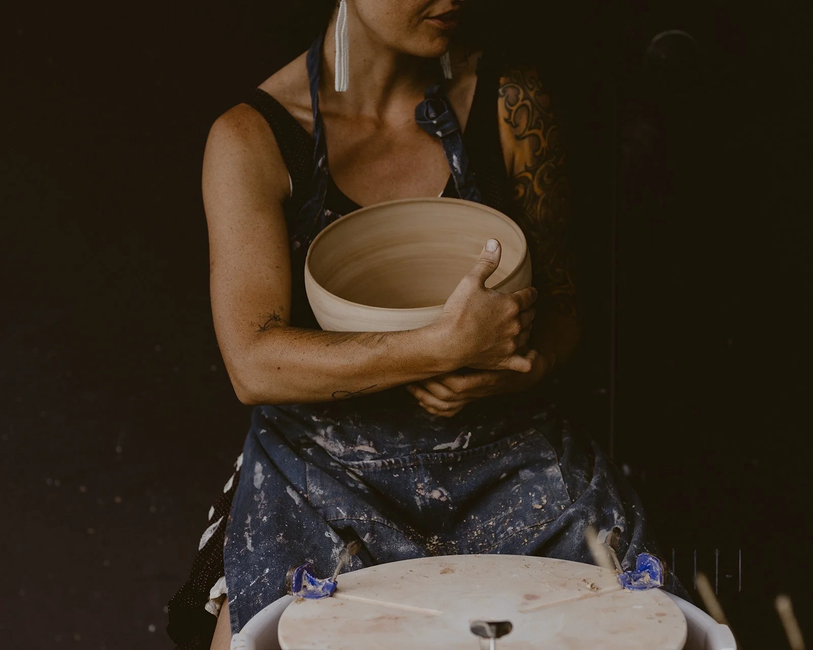 a maker wearing an apron holding a clay bowl, sitting at a pottery wheel.