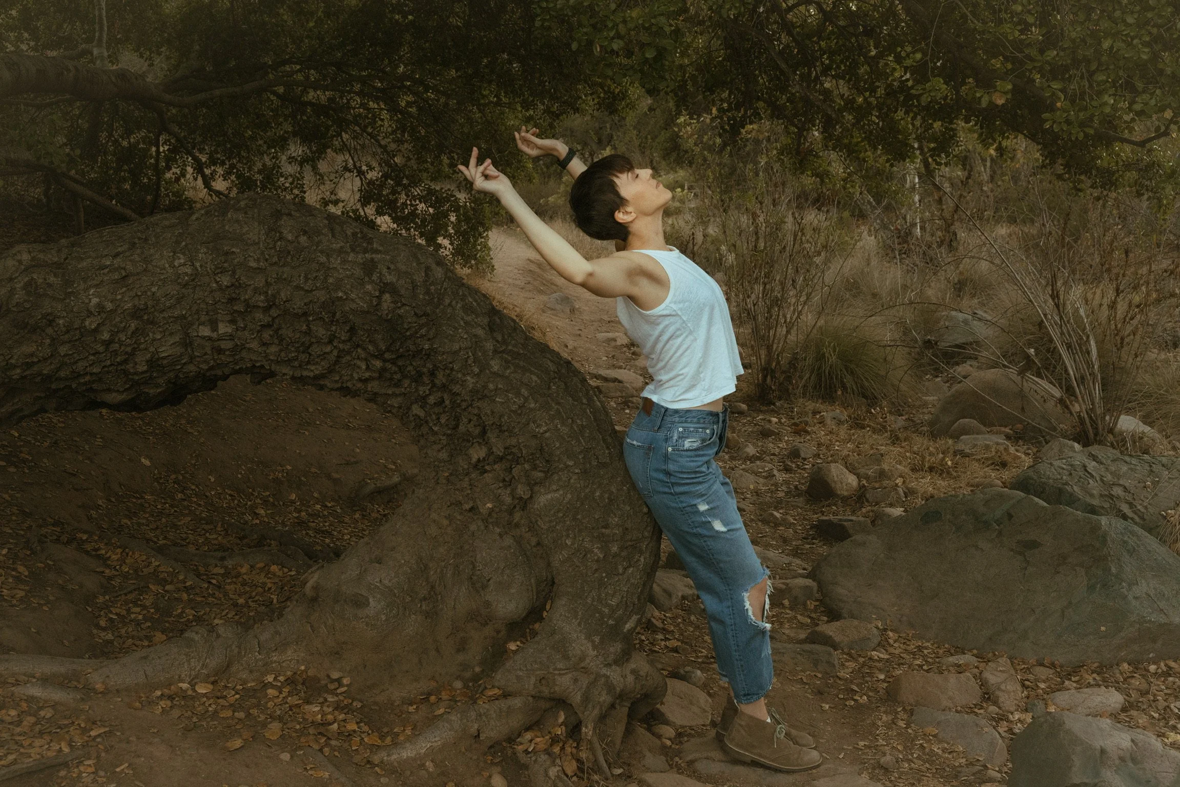 Person in white tank top and ripped jeans leaning back against a large tree branch in a forest setting.