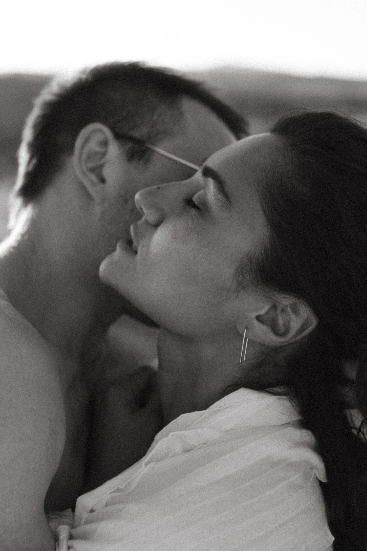A black and white portrait of a couple embracing. The woman has her eyes closed and is wearing a light shirt and an earring. The man's face is partially visible as he hugs her from behind.