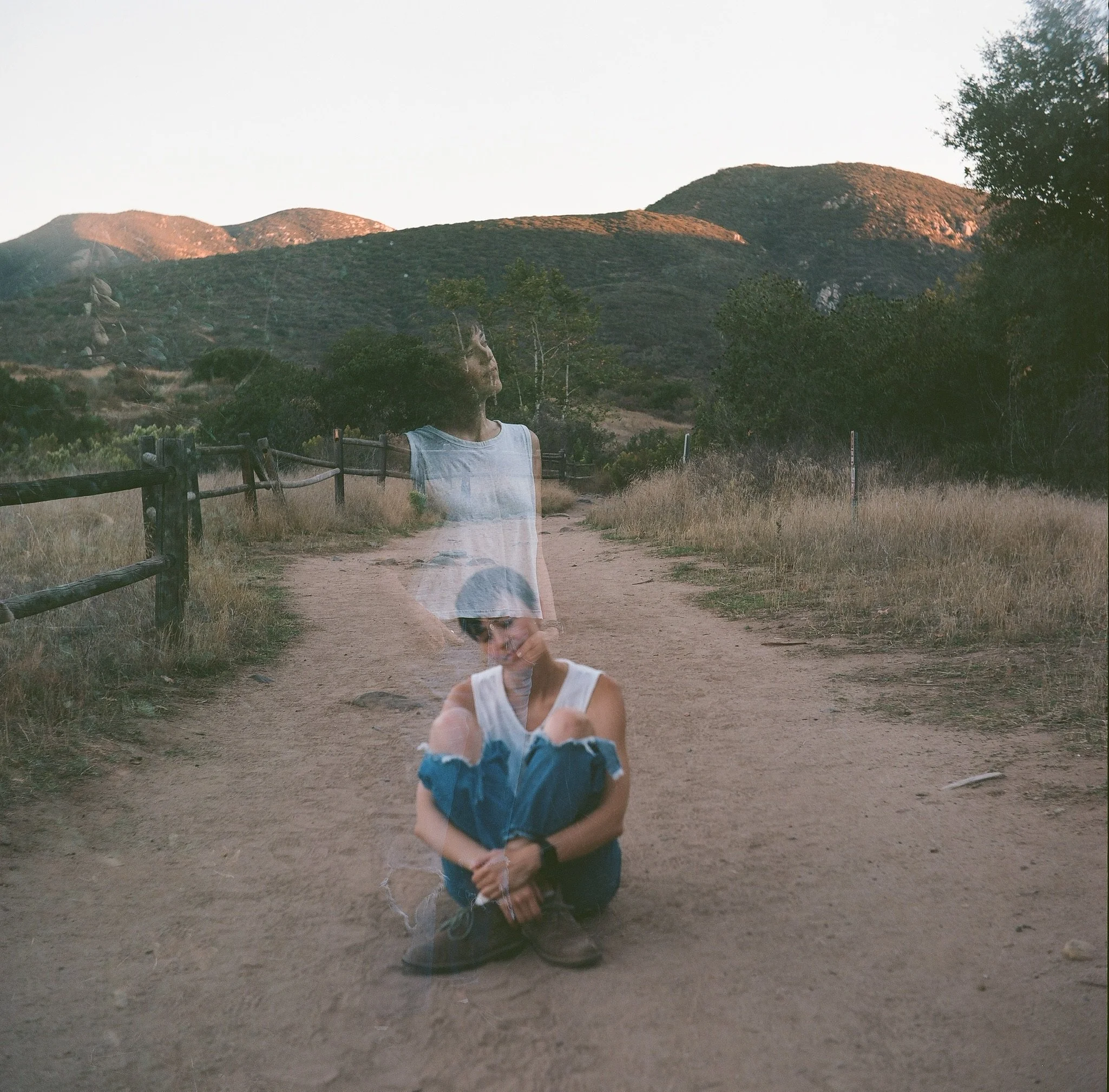 Double exposure photo of a person on a dirt path in a hilly landscape, combining sitting and standing poses, with wooden fencing and dry grass on either side.